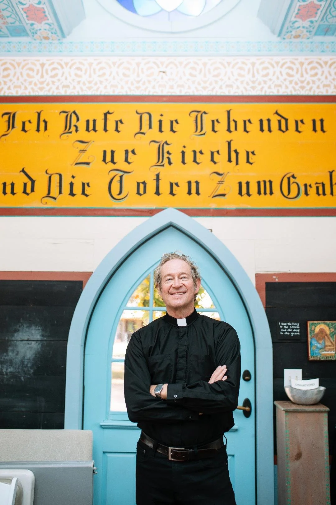 A smiling man dressed as a priest standing with arms crossed in front of a blue church door with a pointed arch, inside a church with a yellow banner with German text and decorative interior.