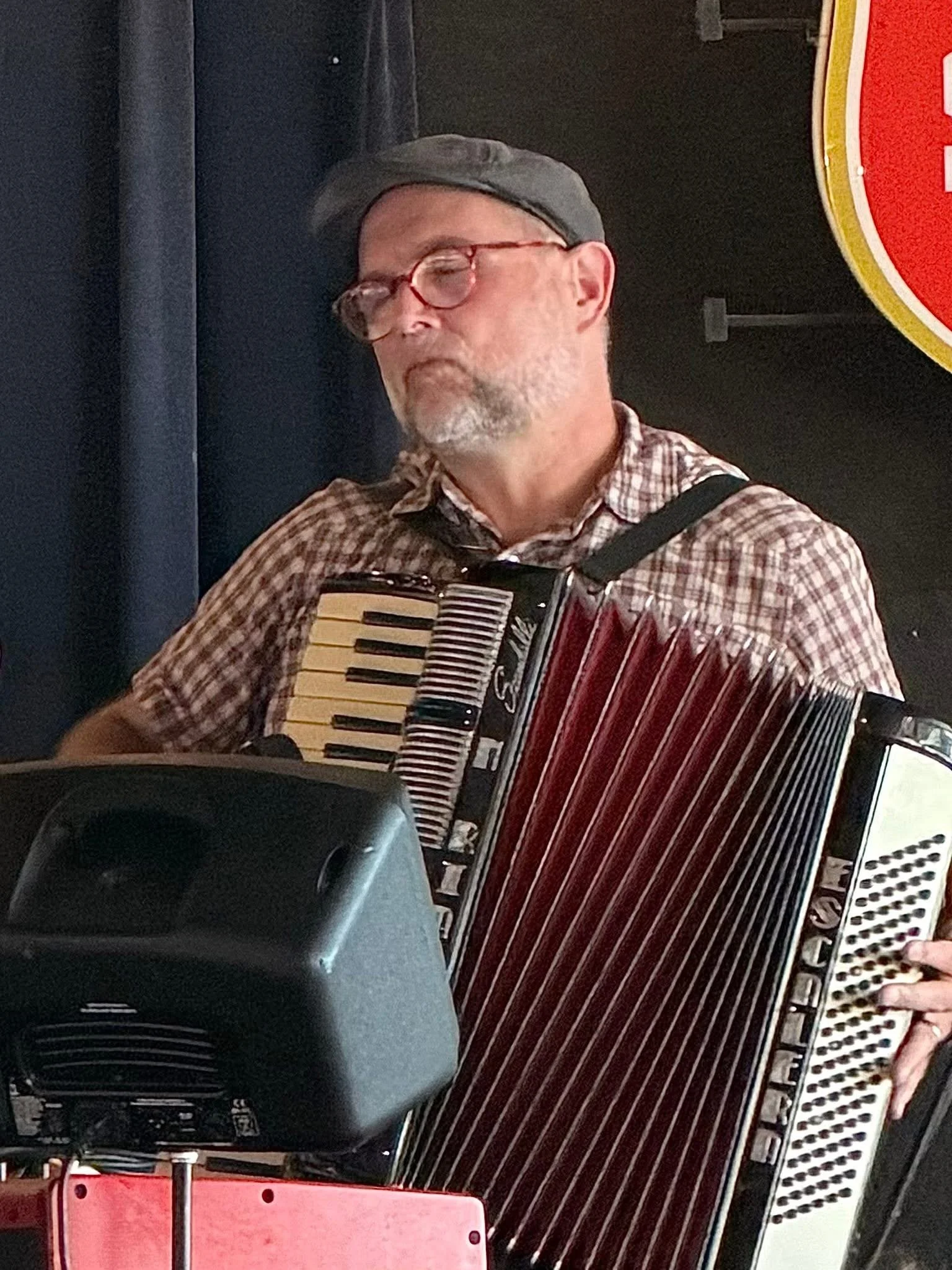 A person wearing glasses, a gray cap, and a plaid shirt is playing an accordion in a dimly lit room with dark walls, with part of a red and white sign visible in the background.
