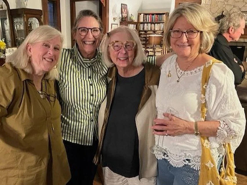 Four women smiling and standing together at a gathering, with a woman in the background at a bar or counter.