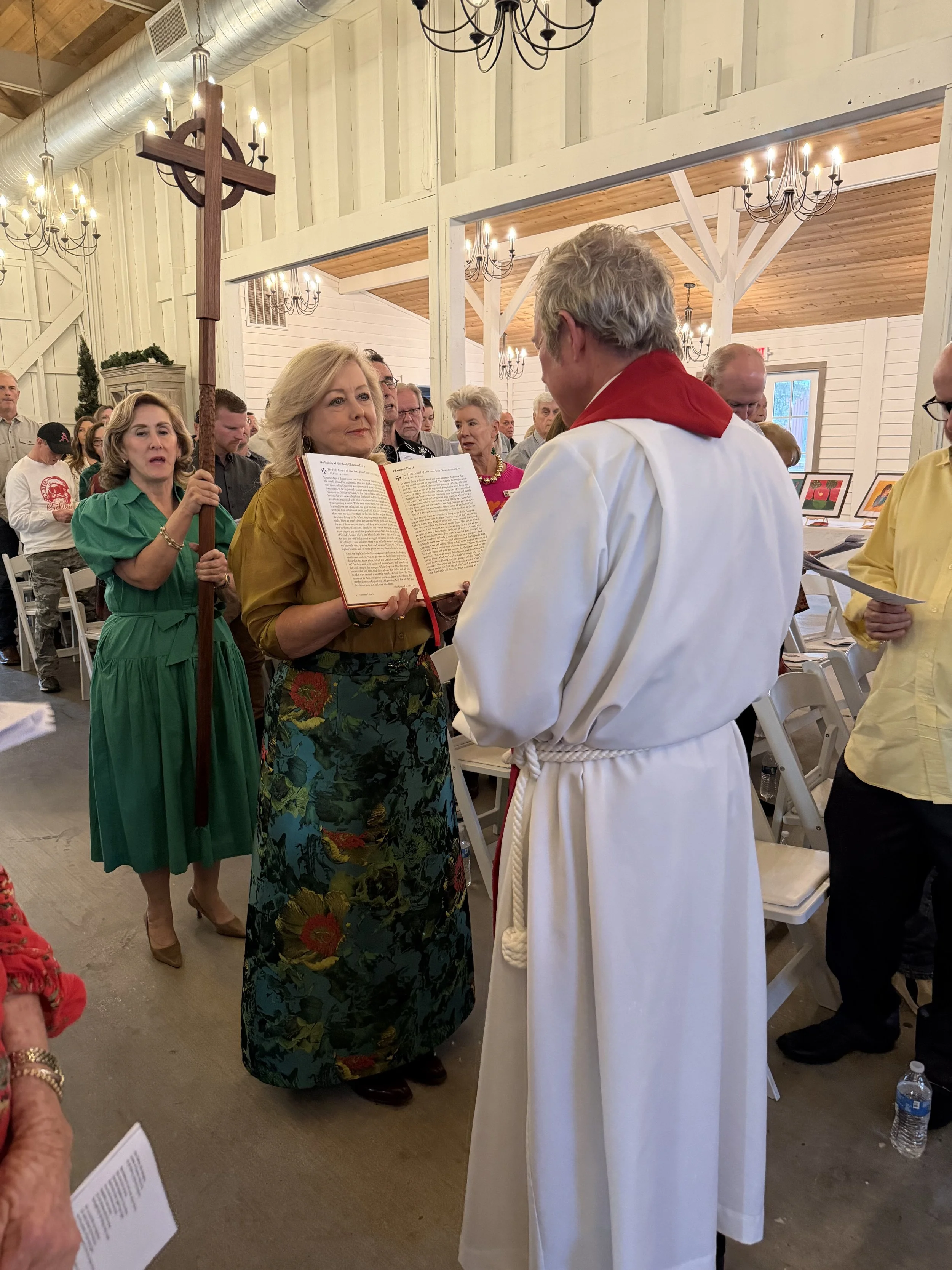A priest in white robes with a red stole officiates a wedding inside a rustic barn with wooden beams, chandeliers, and a cross on a stand. A woman in a mustard blouse and floral skirt holds a book, possibly a Bible, during the ceremony. Guests stand nearby, some holding programs.