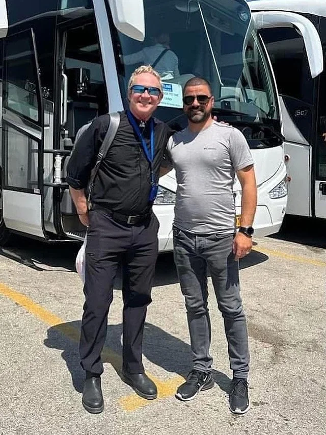 Two men standing in front of a white bus, smiling and posing for the photo. They are outdoors on a sunny day.