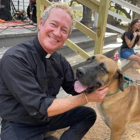 A smiling man in a black shirt crouching next to a large brown dog outdoors, with a woman sitting on steps in the background.