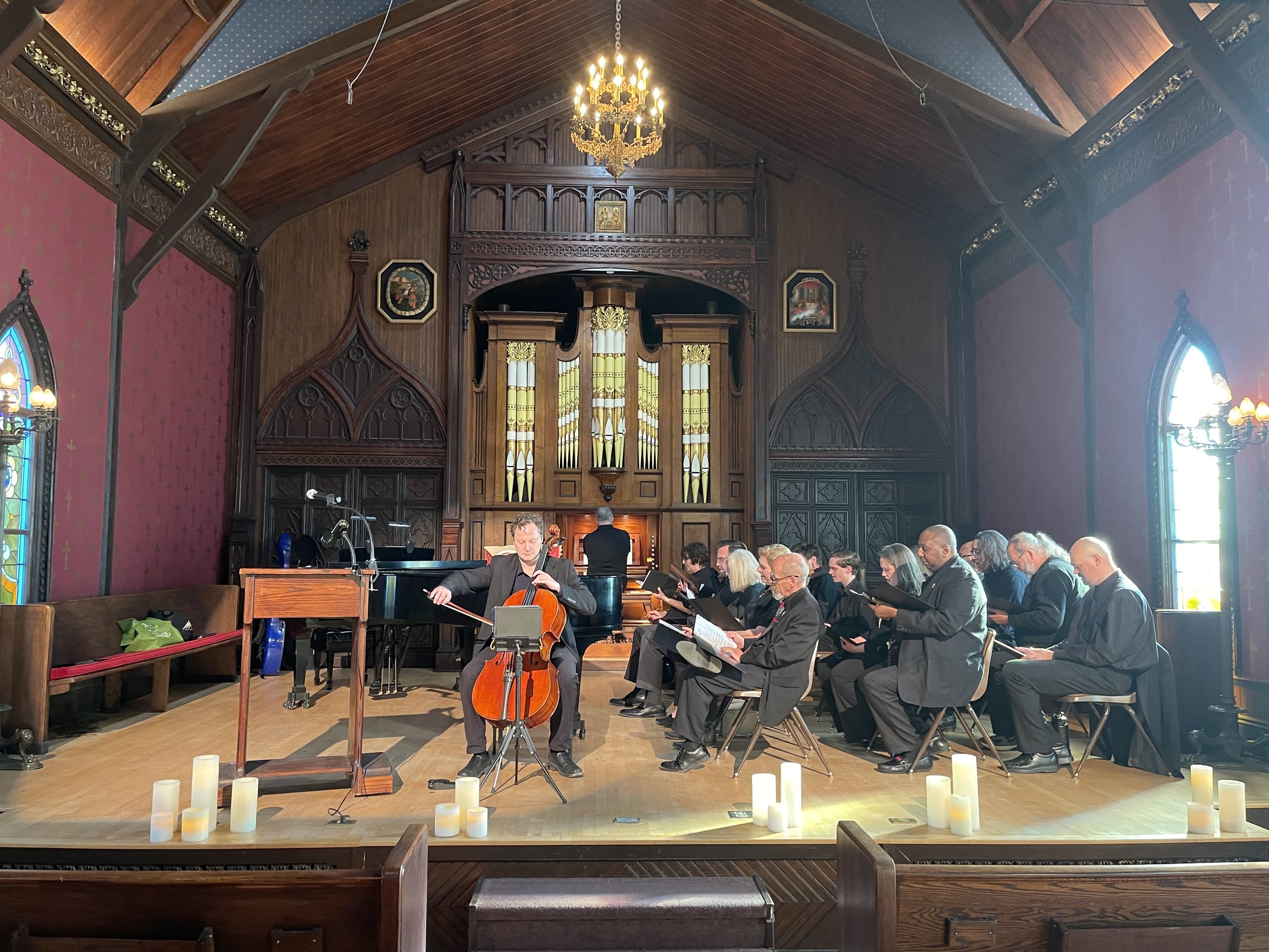 A group of musicians and choir singers performing inside a church with stained glass windows, wooden walls, and a high wooden ceiling. The choir members are seated, reading music sheets, and a cellist is playing in front of them. Candle decorations a