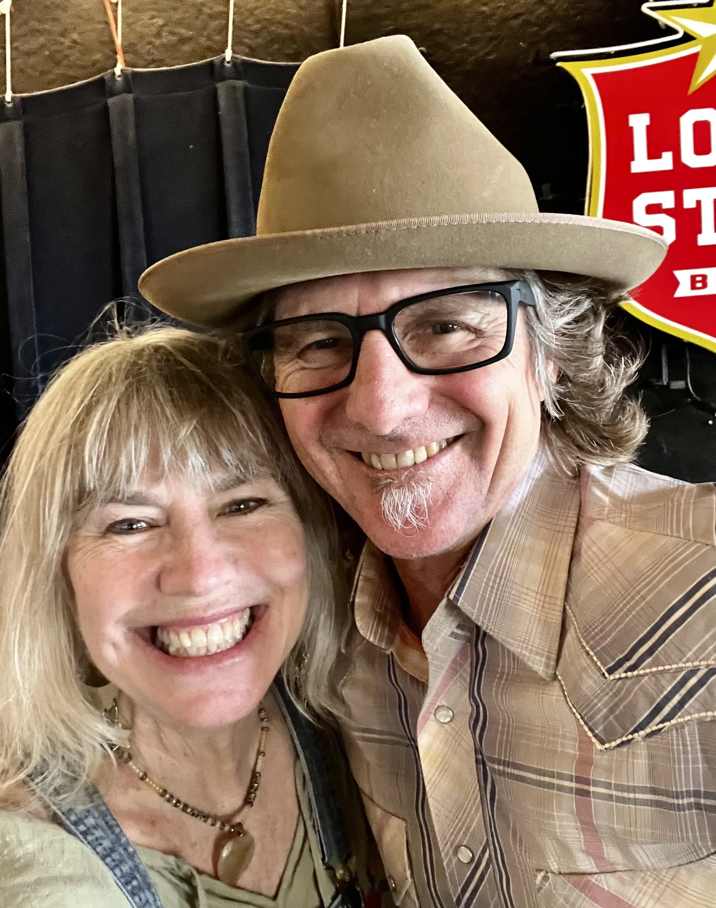 A smiling woman with blonde hair and a necklace, and a smiling man with glasses wearing a tan cowboy hat and a tan plaid shirt, posing together for a selfie.