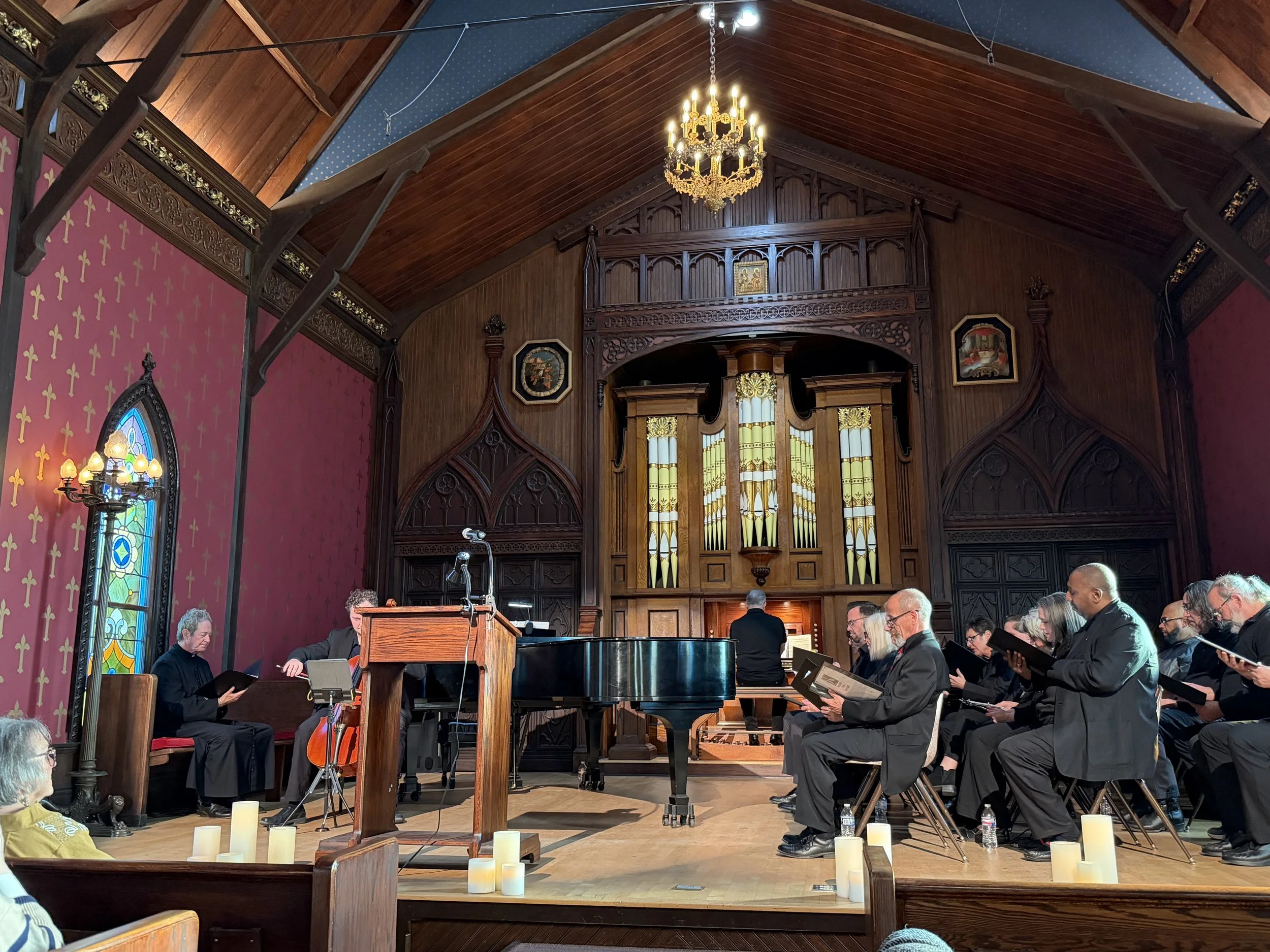 A choir of people singing or rehearsing in a church or concert hall, with a grand pipe organ in the background, candles on the floor, and a chandelier hanging from the wooden ceiling.