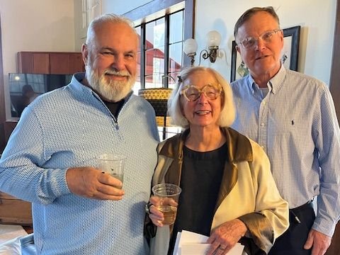 Three older adults, two men and one woman, standing indoors at a social gathering, holding drinks and smiling at the camera.
