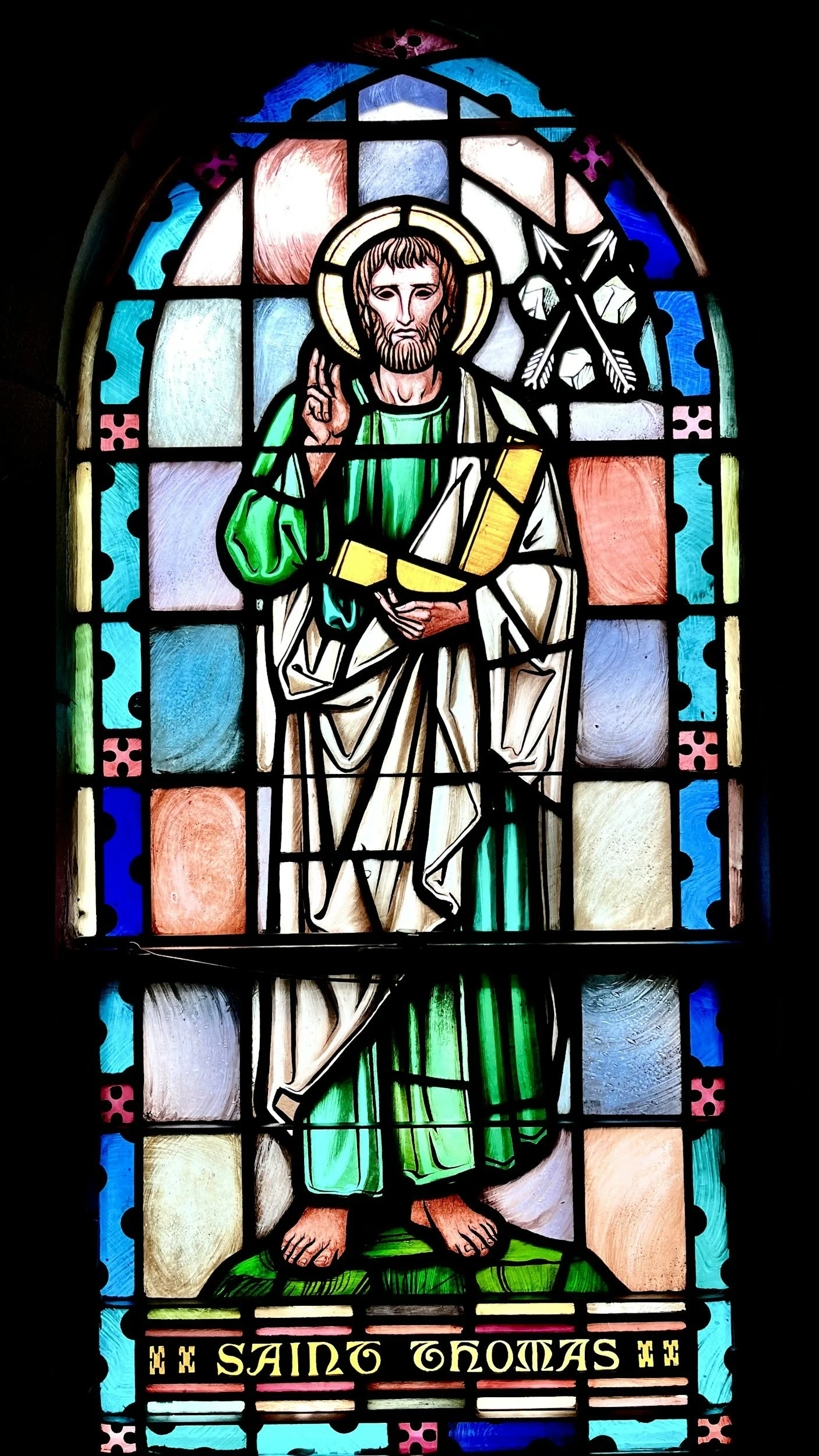 Colorful stained glass window depicting Saint Thomas with a halo, holding a spear and a builder's square, with the words "Saint Thomas" at the bottom.
