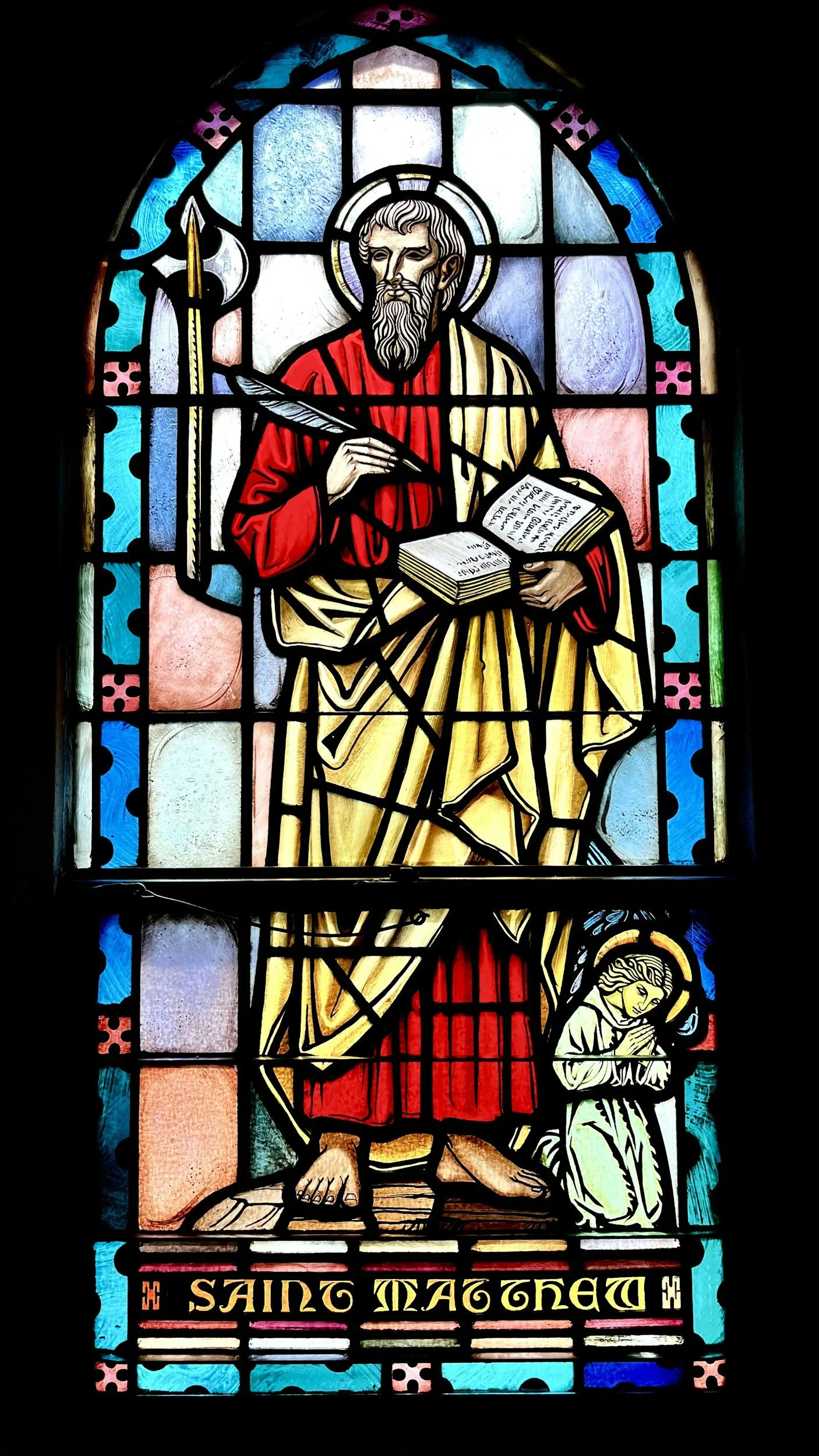 Stained glass window depicting a saint holding a book and writing with a quill, with a kneeling figure in front. The phrase 'SAINT MAGNET' is at the bottom.