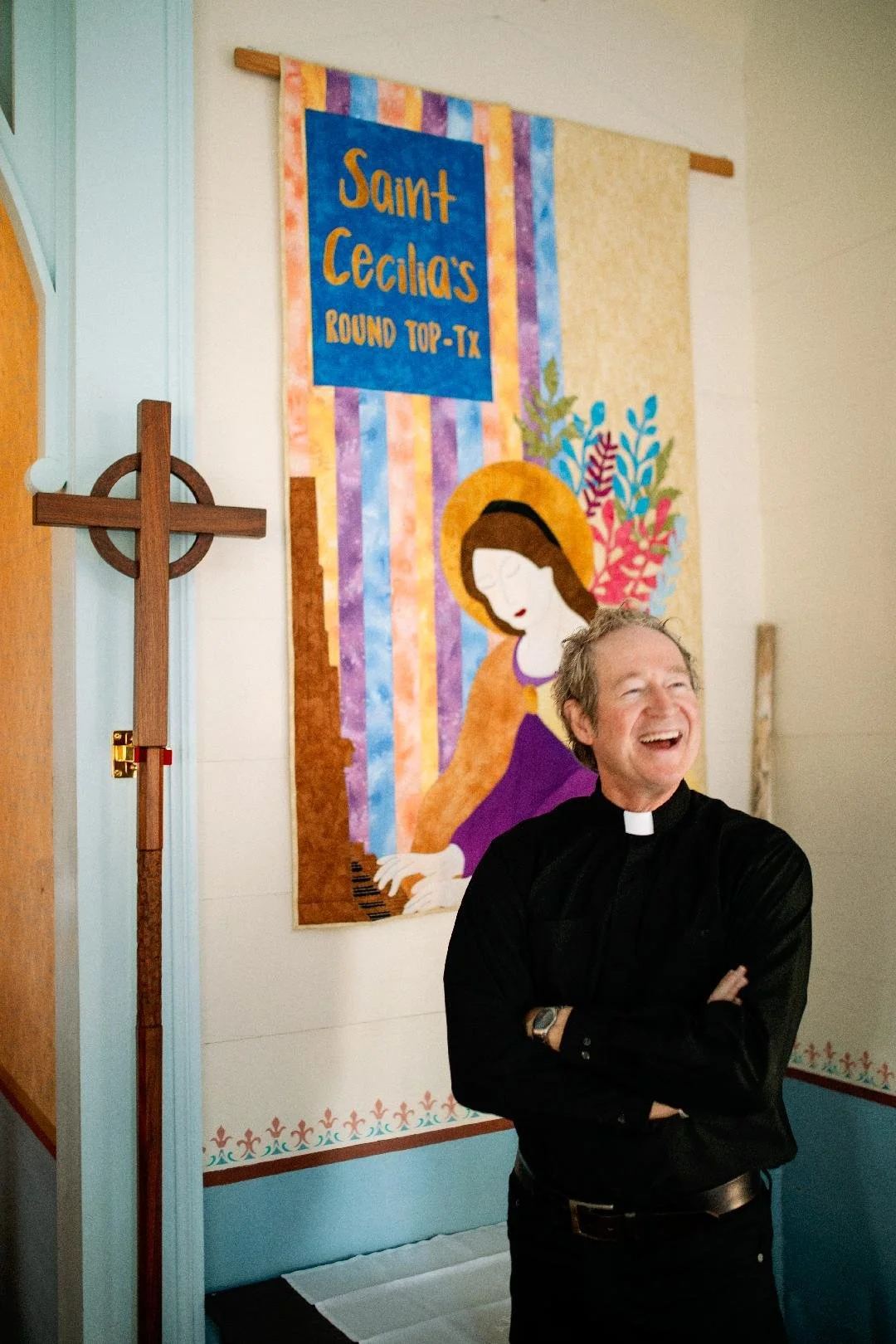 A smiling man dressed as a priest standing in front of a colorful religious banner that depicts a woman playing a piano, with the text 'Saint Cecilia's Round Top, TX'. There is a wooden cross mounted on the wall to the left.