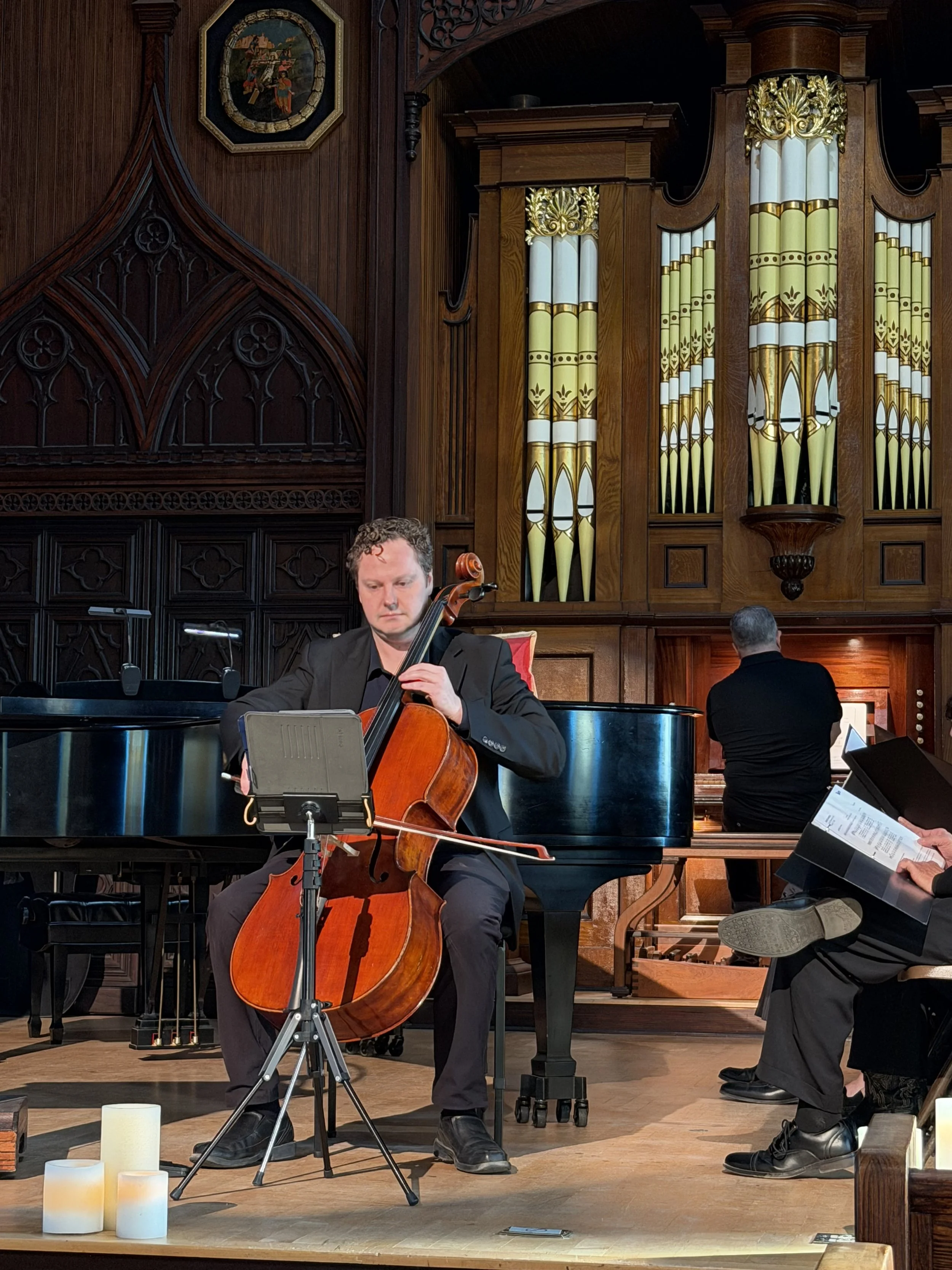Musicians performing in a church or concert hall, with one person playing the cello and another at the piano, and a third holding sheet music.