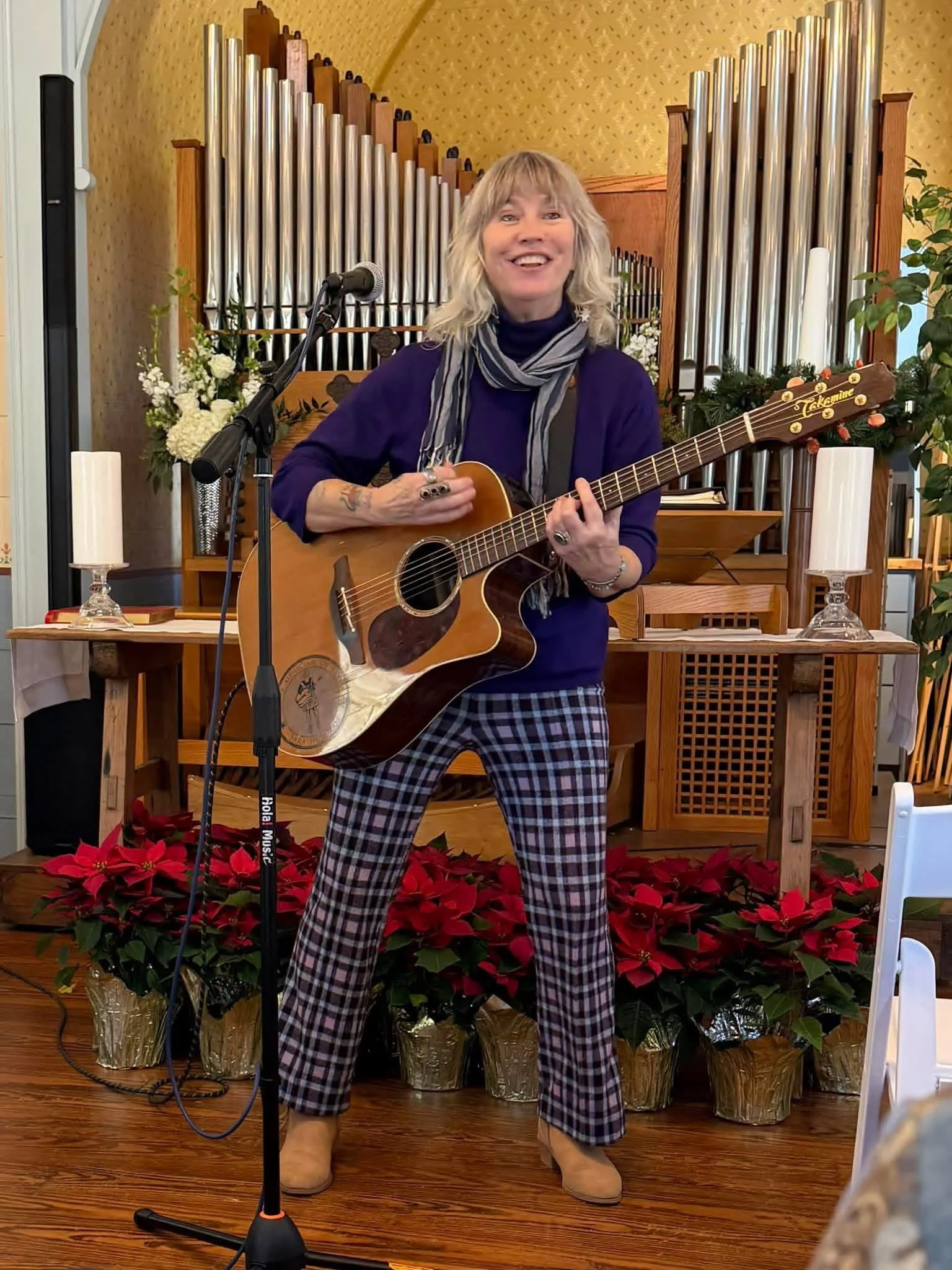 A cheerful woman with shoulder-length blonde hair playing an acoustic guitar on a stage decorated with poinsettias and flowers, with an organ and plants in the background.