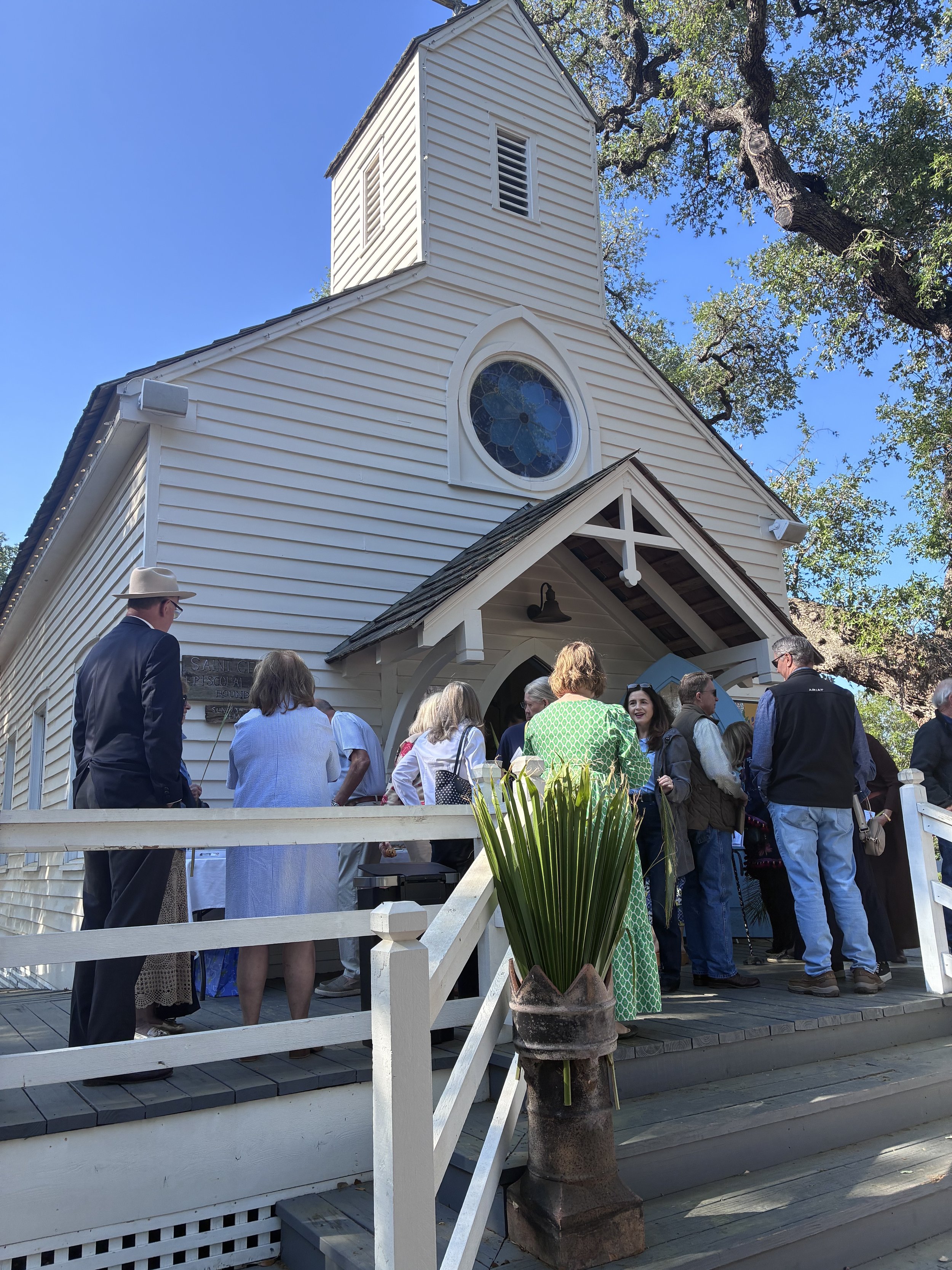 People gathered outside a white church with a bell tower and stained glass window, under a blue sky with trees, during a daytime event or service.