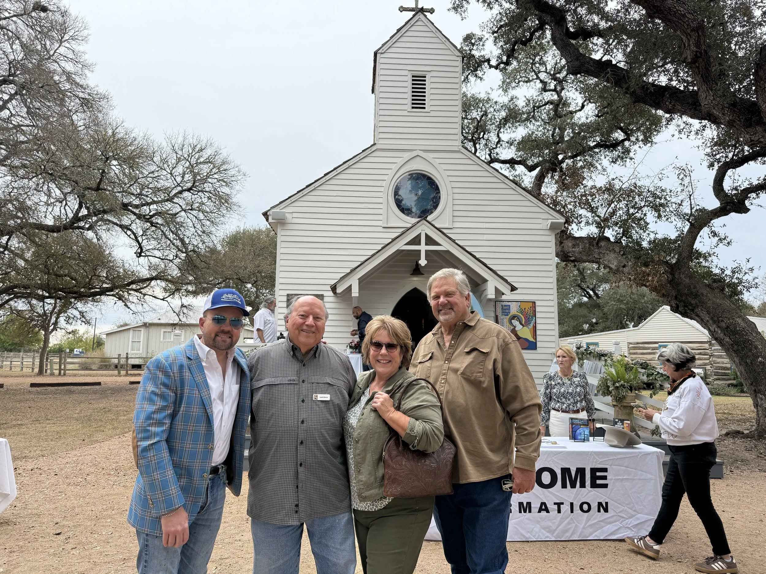 Four people standing in front of a small white church with a steeple and a round stained glass window, outdoors on a cloudy day, with trees and other people visible in the background.