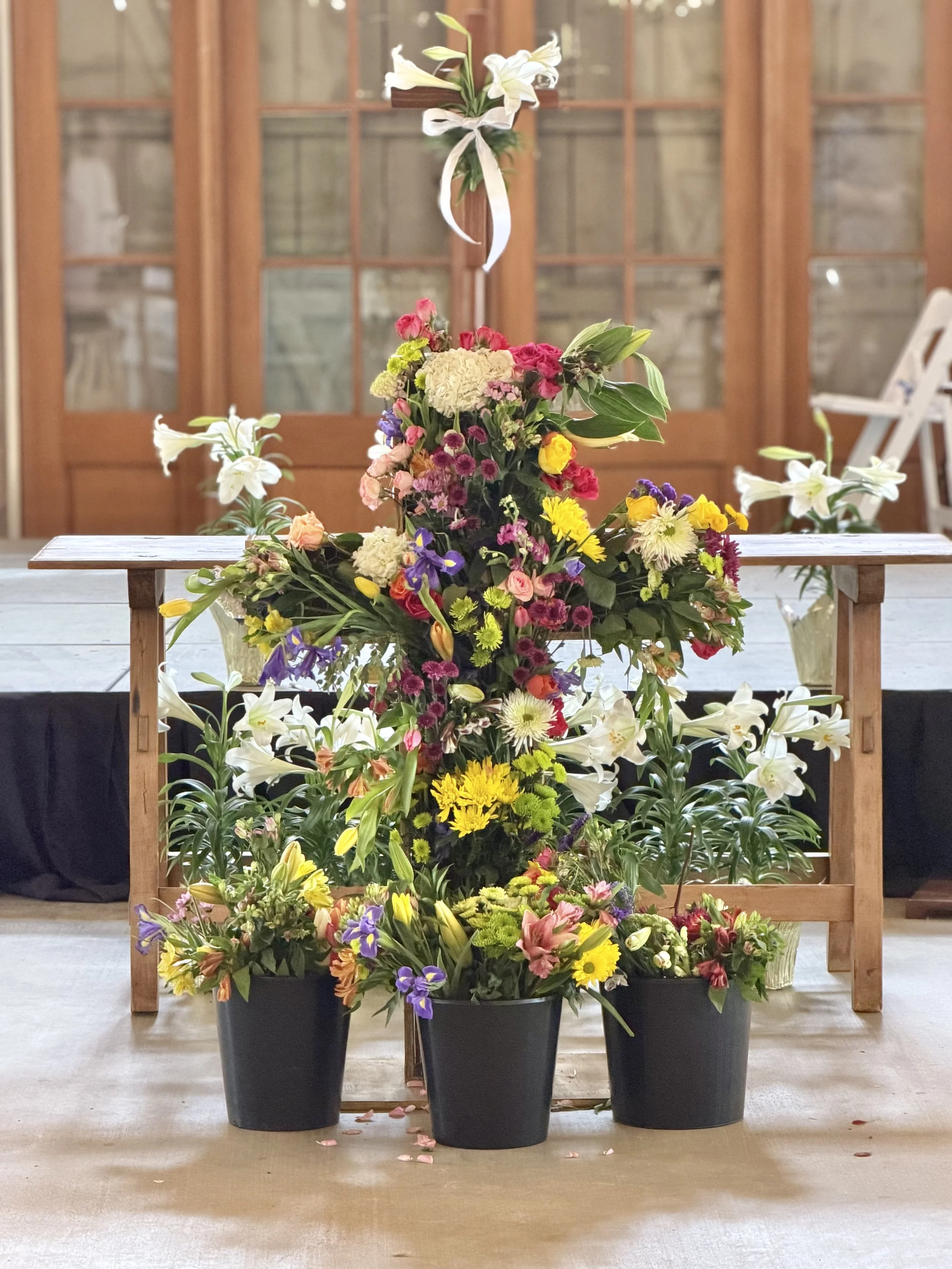 Large arrangement of colorful flowers with white lilies, pink roses, purple, yellow, and white blooms, placed on a wooden table with black pots. Background features a wooden doorway with glass panes.
