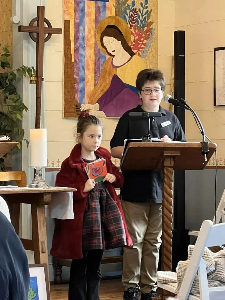 A young boy with glasses and a dark jacket stands at a wooden podium with a microphone, speaking. A young girl with a red coat and plaid dress stands nearby, holding a book. Behind them is a religious painting of Mary and child, and to the side is a 