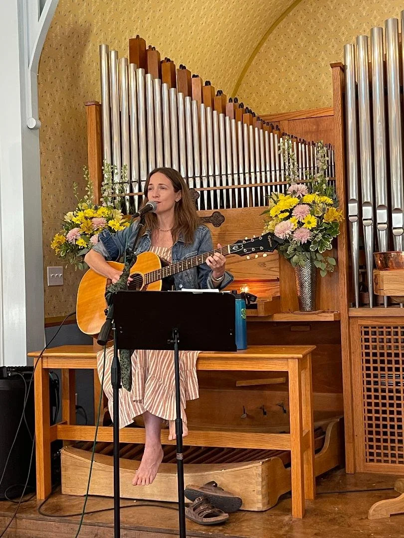 A woman is sitting on a wooden table playing an acoustic guitar and singing into a microphone. She is barefoot with one foot resting on a small platform. Behind her is a large pipe organ with tall metal pipes and a wooden frame, and there are two lar