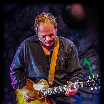 A man playing an electric guitar on stage, wearing a black shirt with a yellow strap, with dark background and stage lighting.