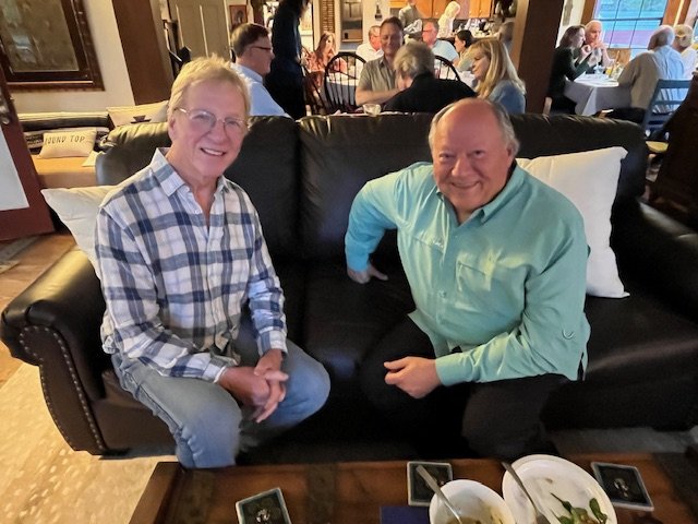 Two men sitting on a black leather couch in a restaurant, smiling at the camera. The man on the left is wearing glasses and a plaid shirt; the man on the right is wearing a teal shirt. There are many people dining in the background.