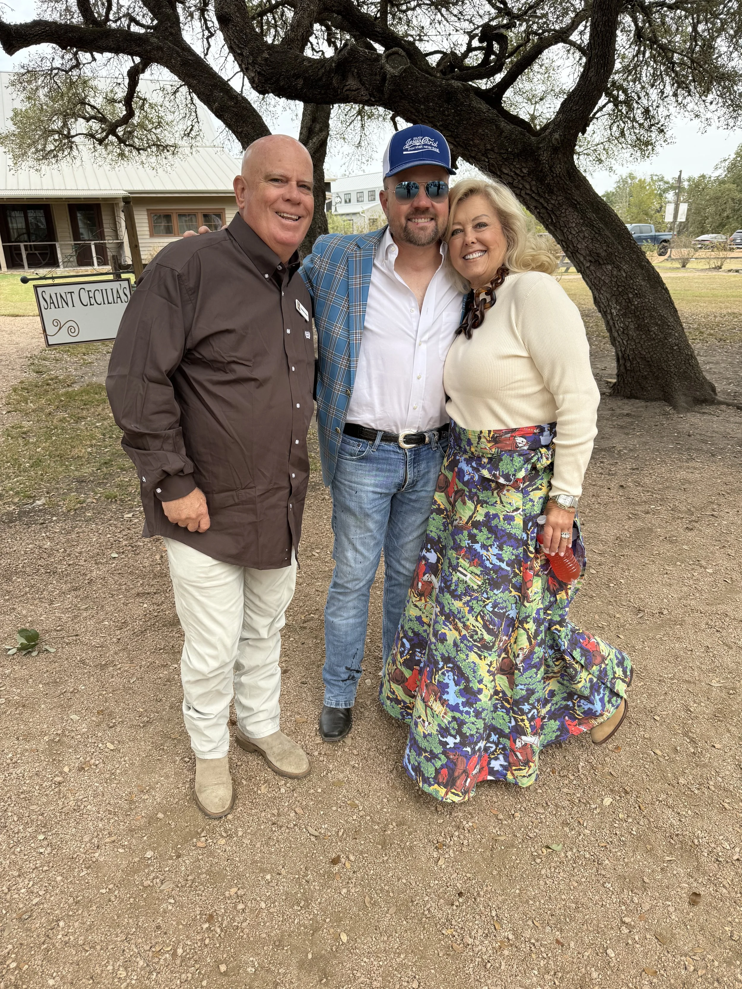 Three people standing outdoors with a large tree behind them, smiling, with a house and parking lot in the background.
