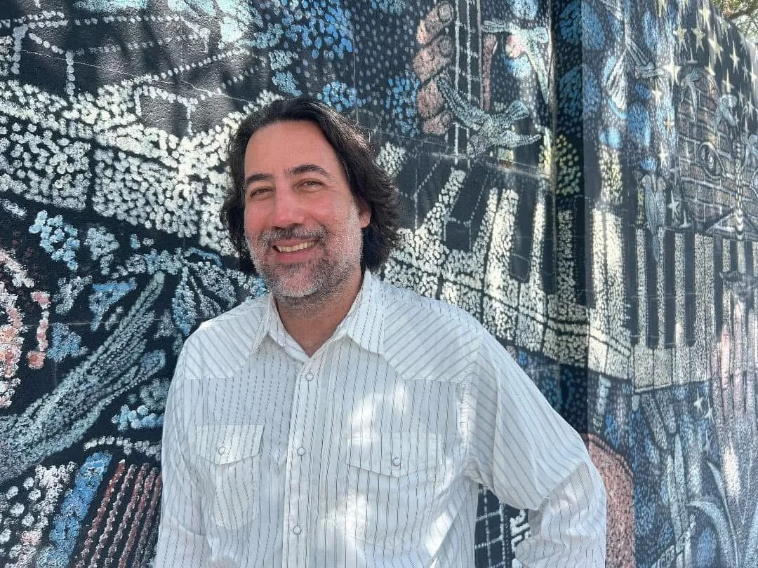 A smiling man with dark shoulder-length hair and a beard, wearing a white striped shirt, standing in front of a large mural with various detailed designs and patterns.