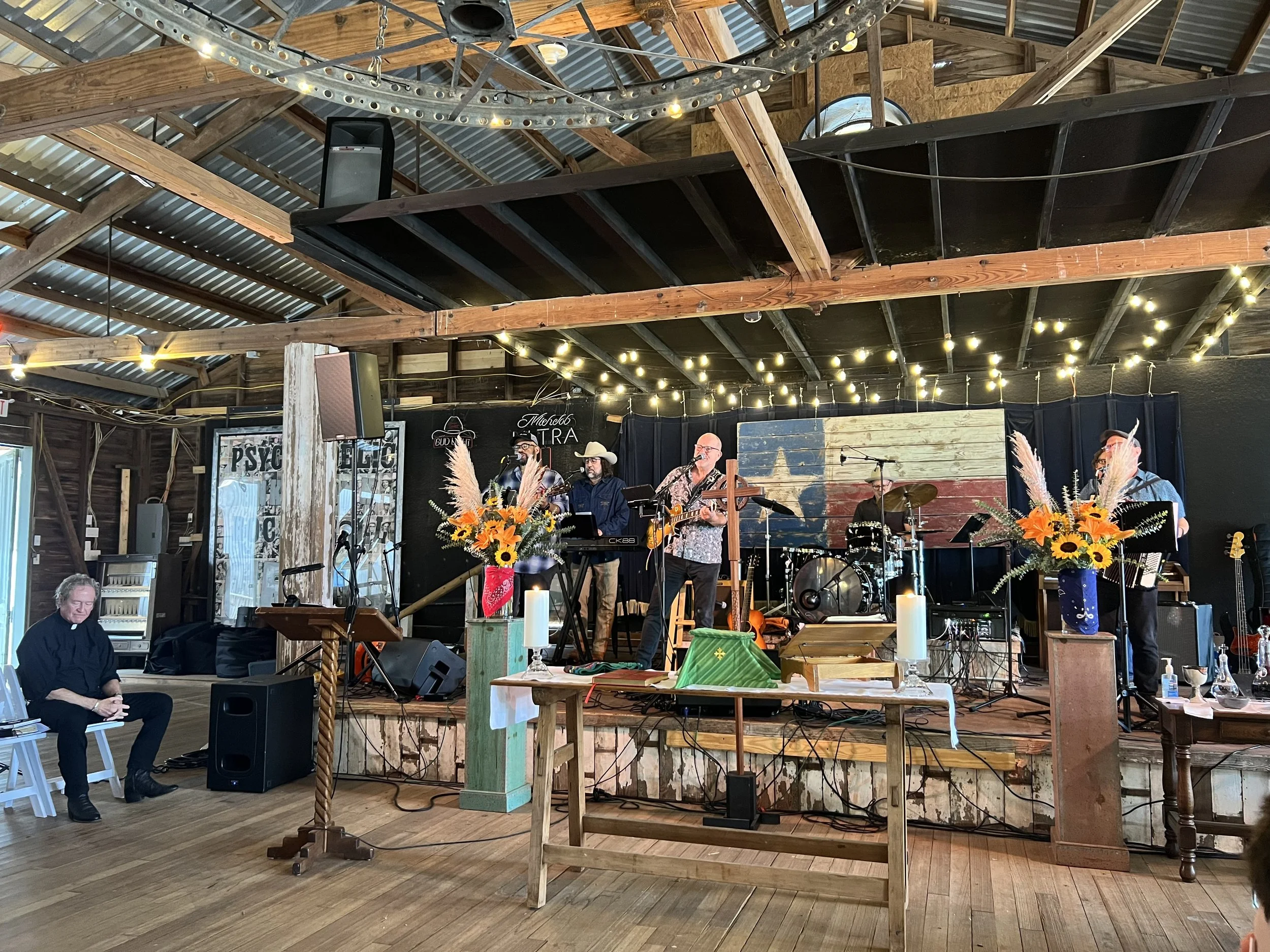 A band performing on stage inside a rustic venue with wooden beams and string lights overhead. There are musicians playing instruments, and flower arrangements in front of the stage.