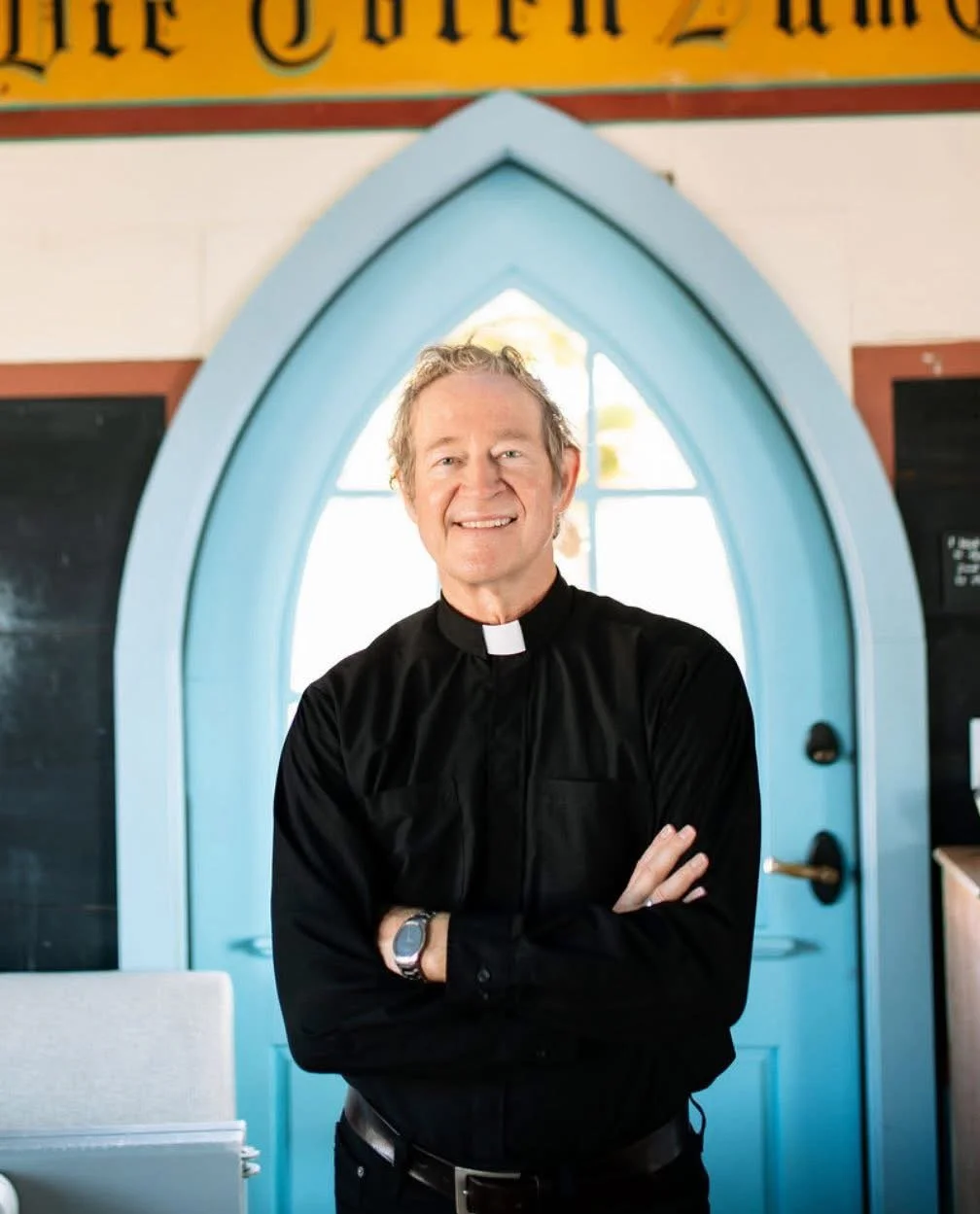A smiling man dressed as a priest in a black clerical shirt with a white collar, standing with his arms crossed in front of a bright church window with an arched shape and light blue trim.