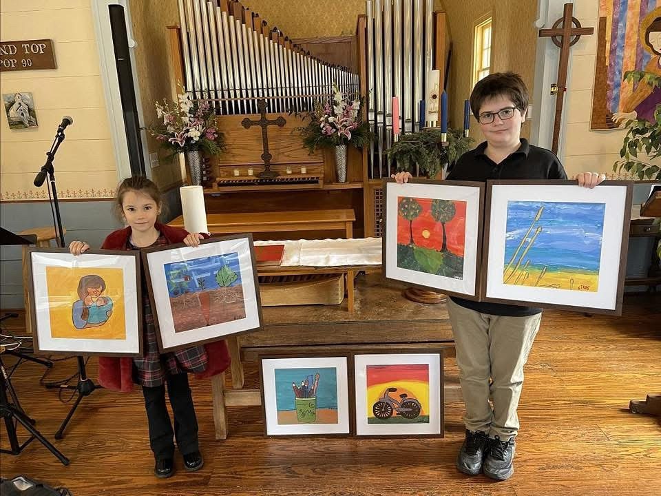 Two children holding framed artwork in a church setting, with more paintings displayed on the table and a wooden organ in the background.