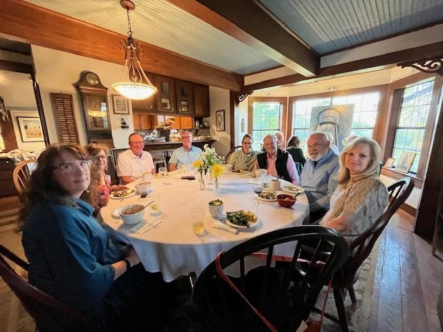 Family gathering around a dining table in a cozy, well-lit room with large windows, enjoying a meal together.