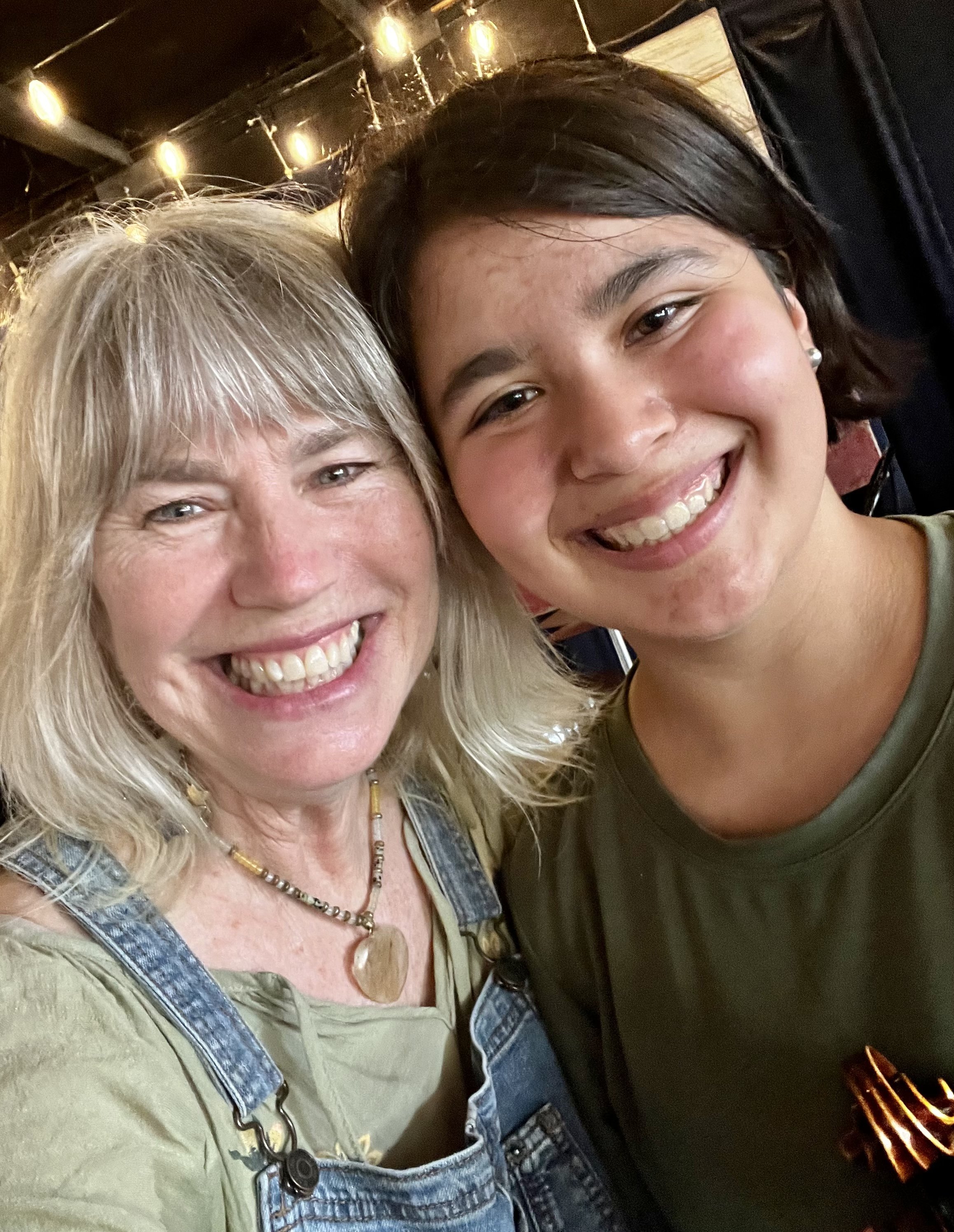 A smiling older woman with blonde hair wearing a denim jumper over a green top, and a necklace with a heart pendant, standing next to a smiling young woman with short dark hair, earrings, and a black top. They are close together and smiling for the c