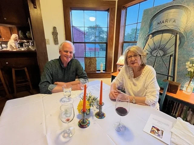 Two older adults, a man and a woman, sitting at a dining table with white tablecloth, glasses of water and red wine, and decorative orange candles in holders. The woman appears to be speaking, the man is smiling. The background shows windows with tre