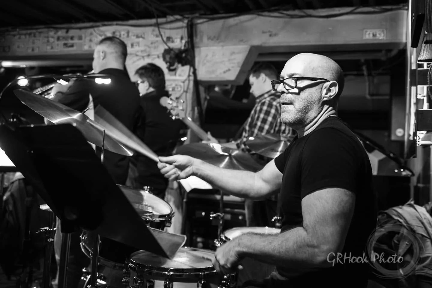 A black-and-white photo of a man playing drums, wearing glasses and a black t-shirt, with other musicians in the background.