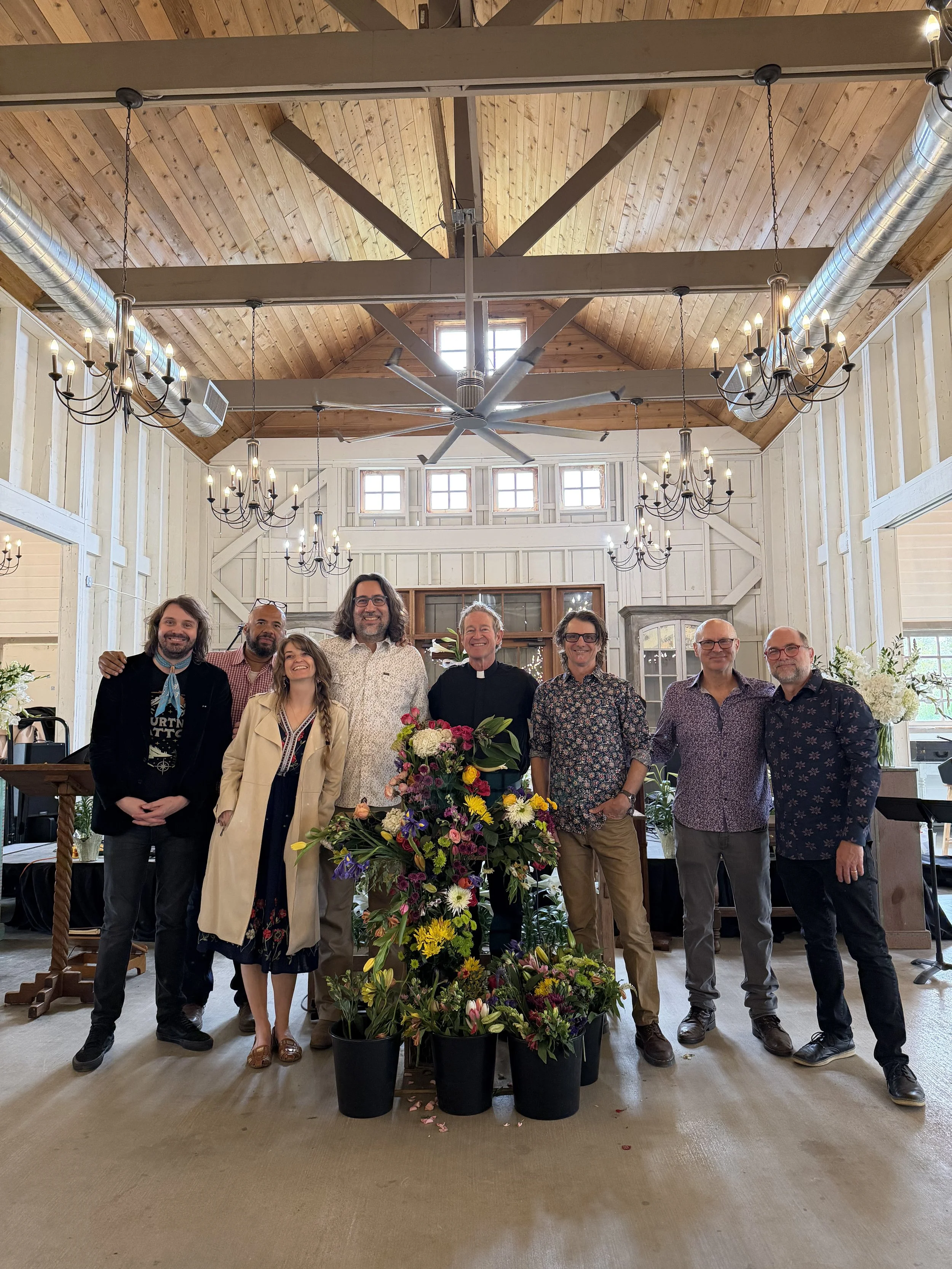 A group of ten people standing together in front of a table with colorful flowers, inside a building with a wooden ceiling and chandeliers.