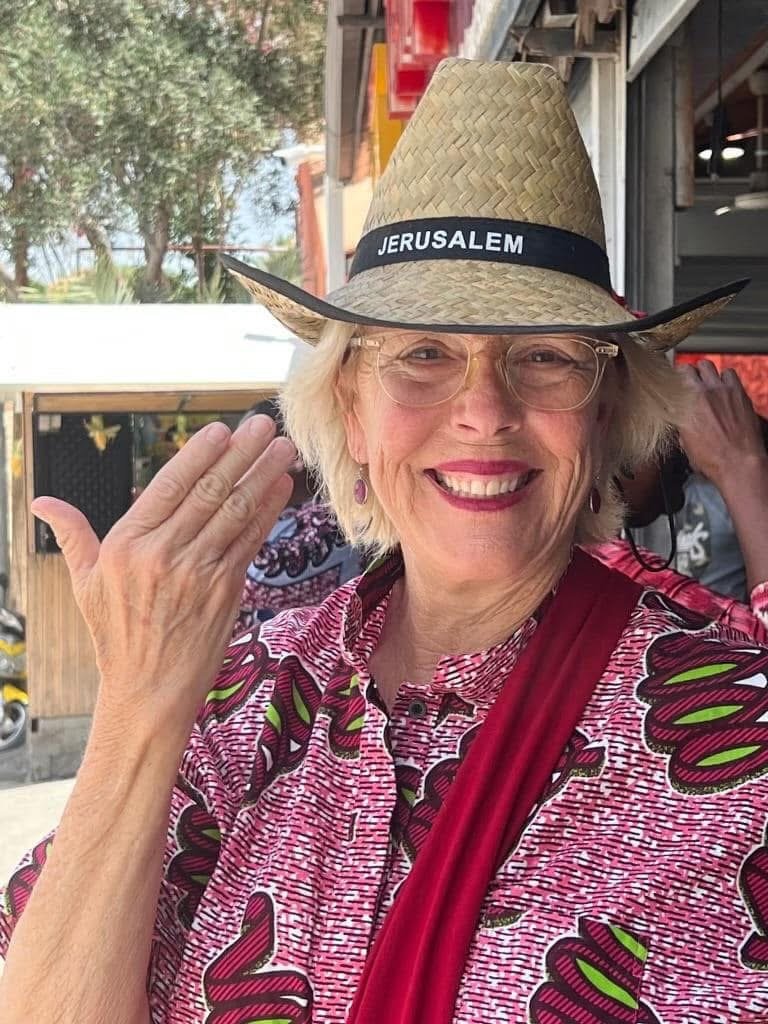 A smiling older woman wearing glasses and a patterned pink shirt, with a red strap across her chest, is smiling and waving with her right hand. She is wearing a straw hat labeled "Jerusalem."