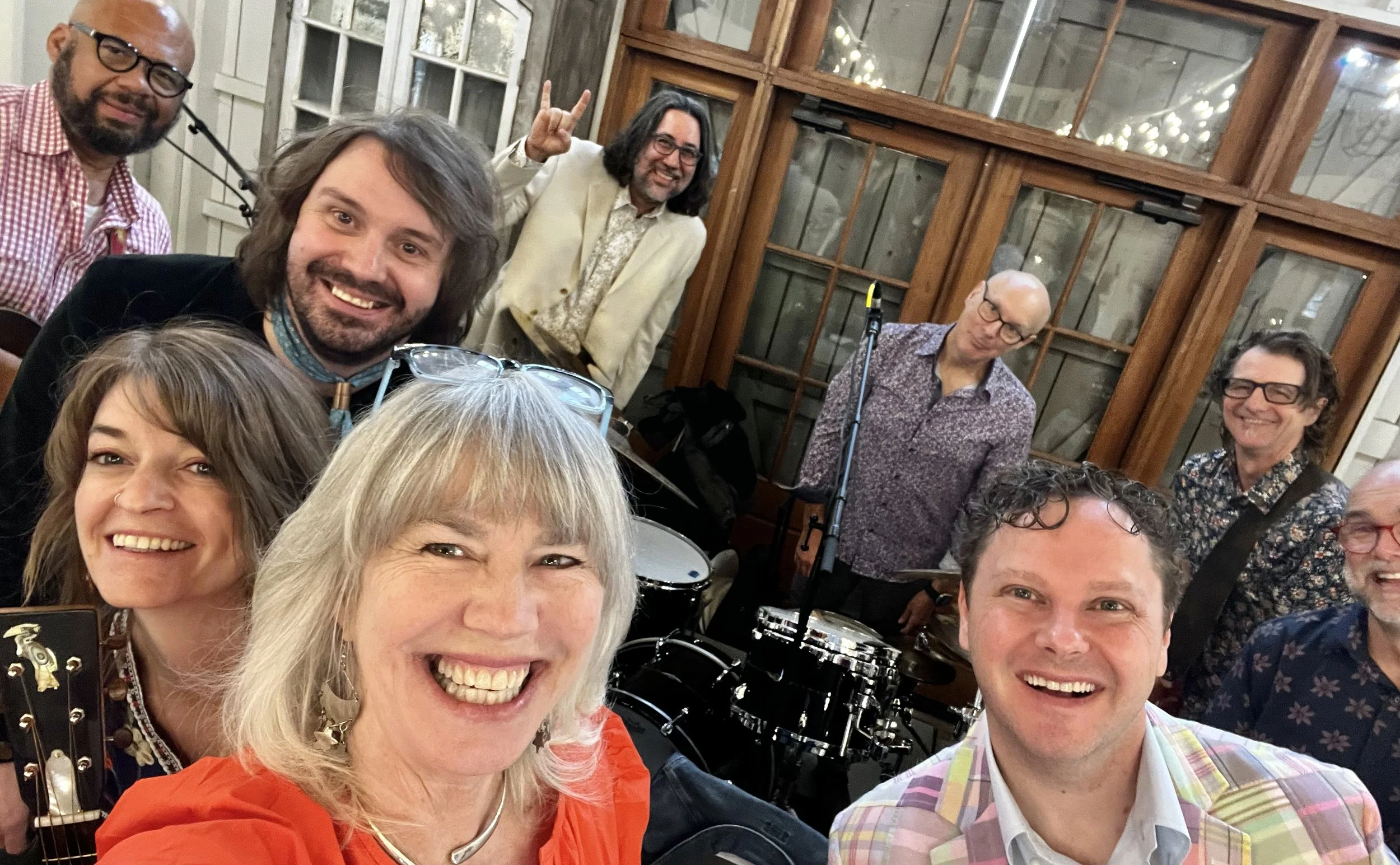 A group of smiling people, some holding musical instruments, are gathered together in a warmly lit room with wooden-framed windows in the background.