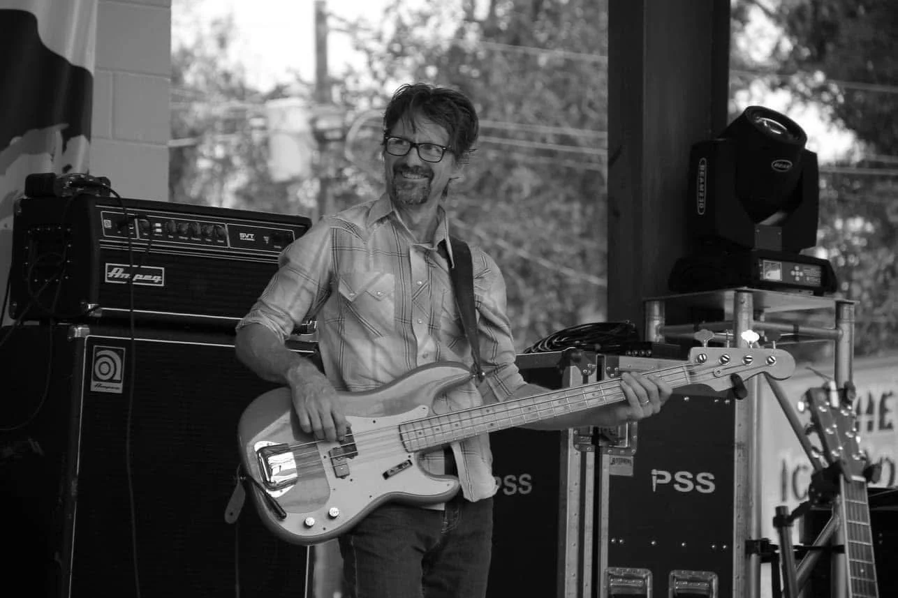A man with glasses and a beard playing a bass guitar on stage, smiling, with audio equipment and trees visible in the background.