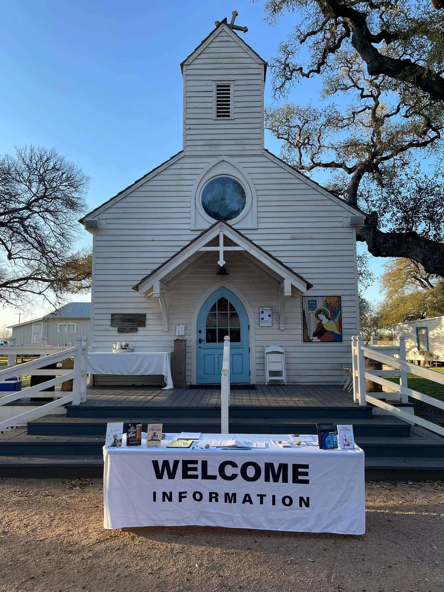 White church with a blue door and a circular window, set against a clear sky with tree branches. In front, a table with informational materials and a large sign that says "WELCOME INFORMATION."