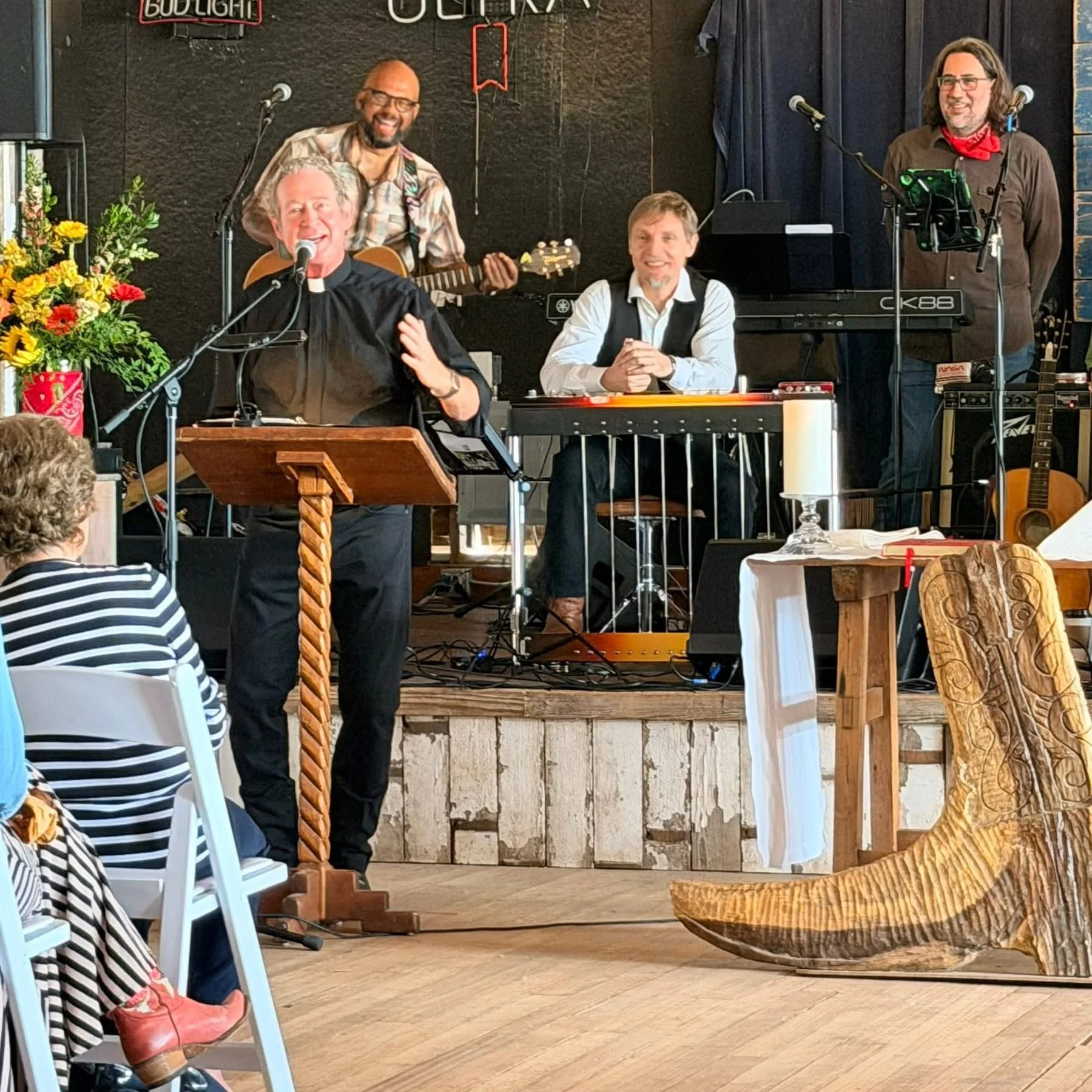 Four men performing on a stage in a restaurant or cafe, with microphones and musical instruments. A woman in the audience is seen in the foreground wearing a striped shirt and red shoes. The stage features a black backdrop, a keyboard, and a guitar. 