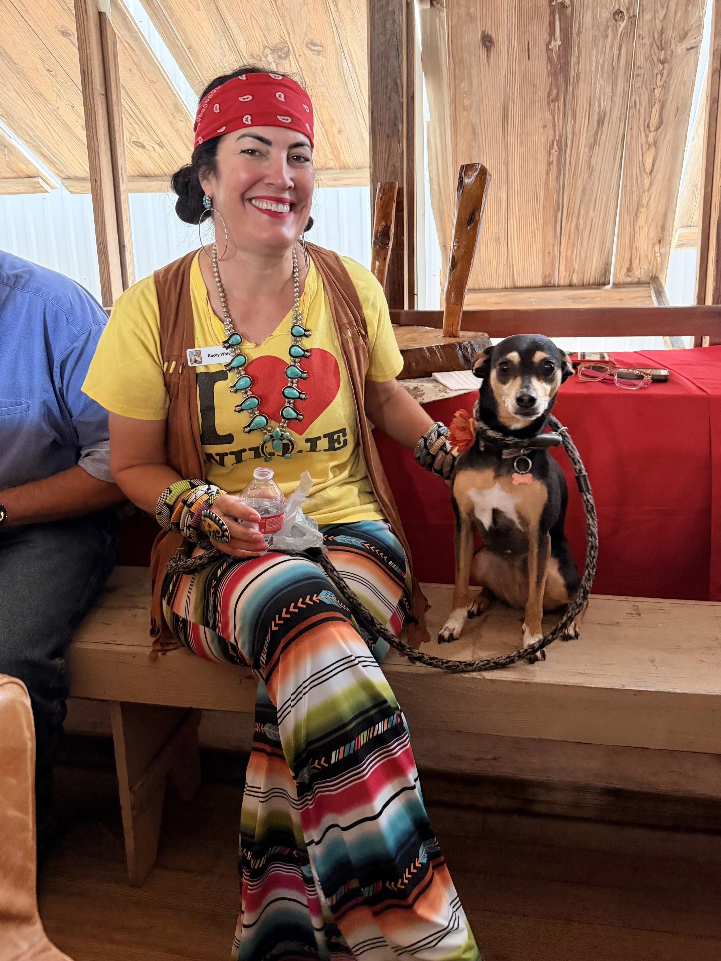 A woman with a red bandana, yellow t-shirt with a red heart and 'LOVE' text, colorful striped pants, and jewelry, sitting indoors with a small black and tan dog.