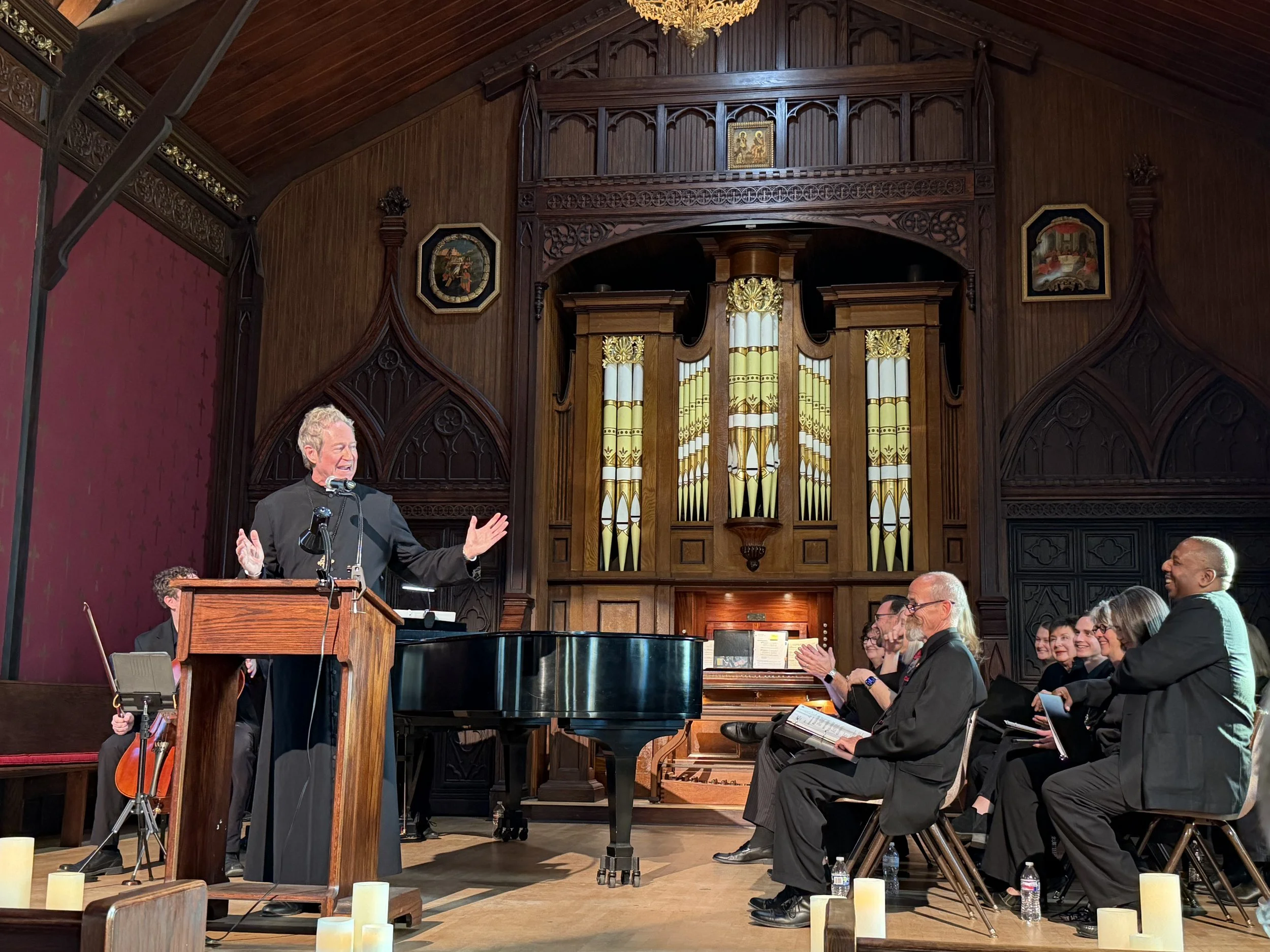 A man in a black suit is speaking at a podium in a large, wood-paneled room with a pipe organ behind him. Several people are seated and clapping, smiling, and listening, while some have notebooks and water bottles.
