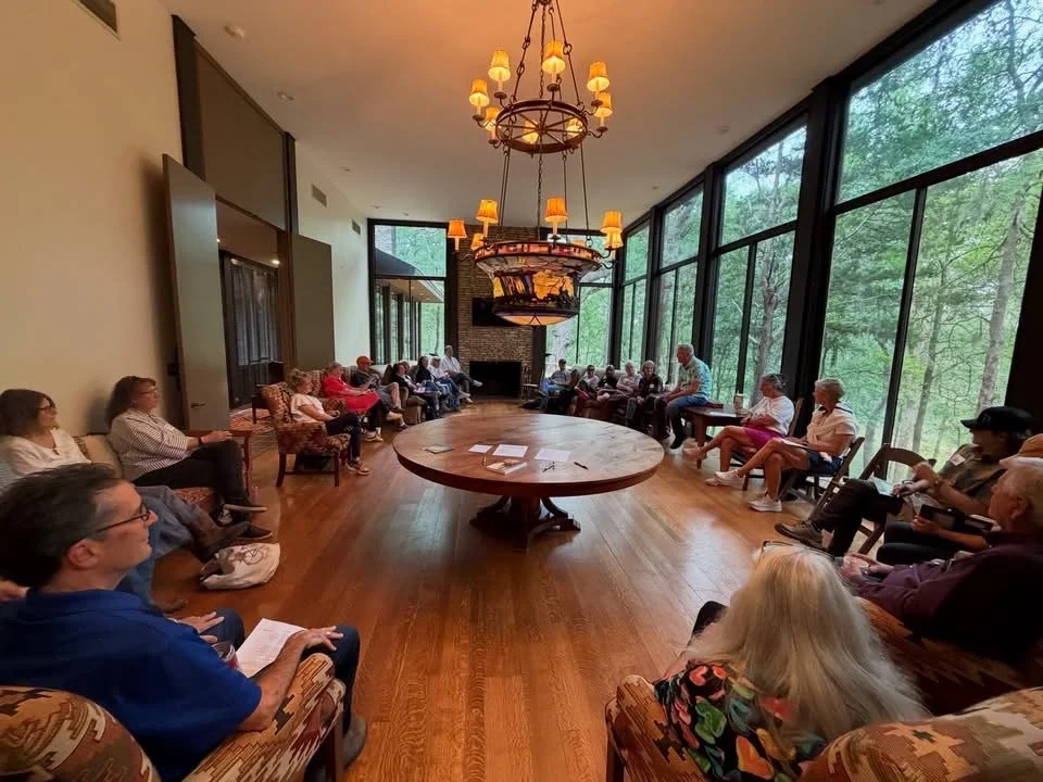 A group of elderly people sitting in a circle in a room with floor-to-ceiling windows showing a forest. There are chandeliers hanging from the ceiling, and a large round table in the center of the room.