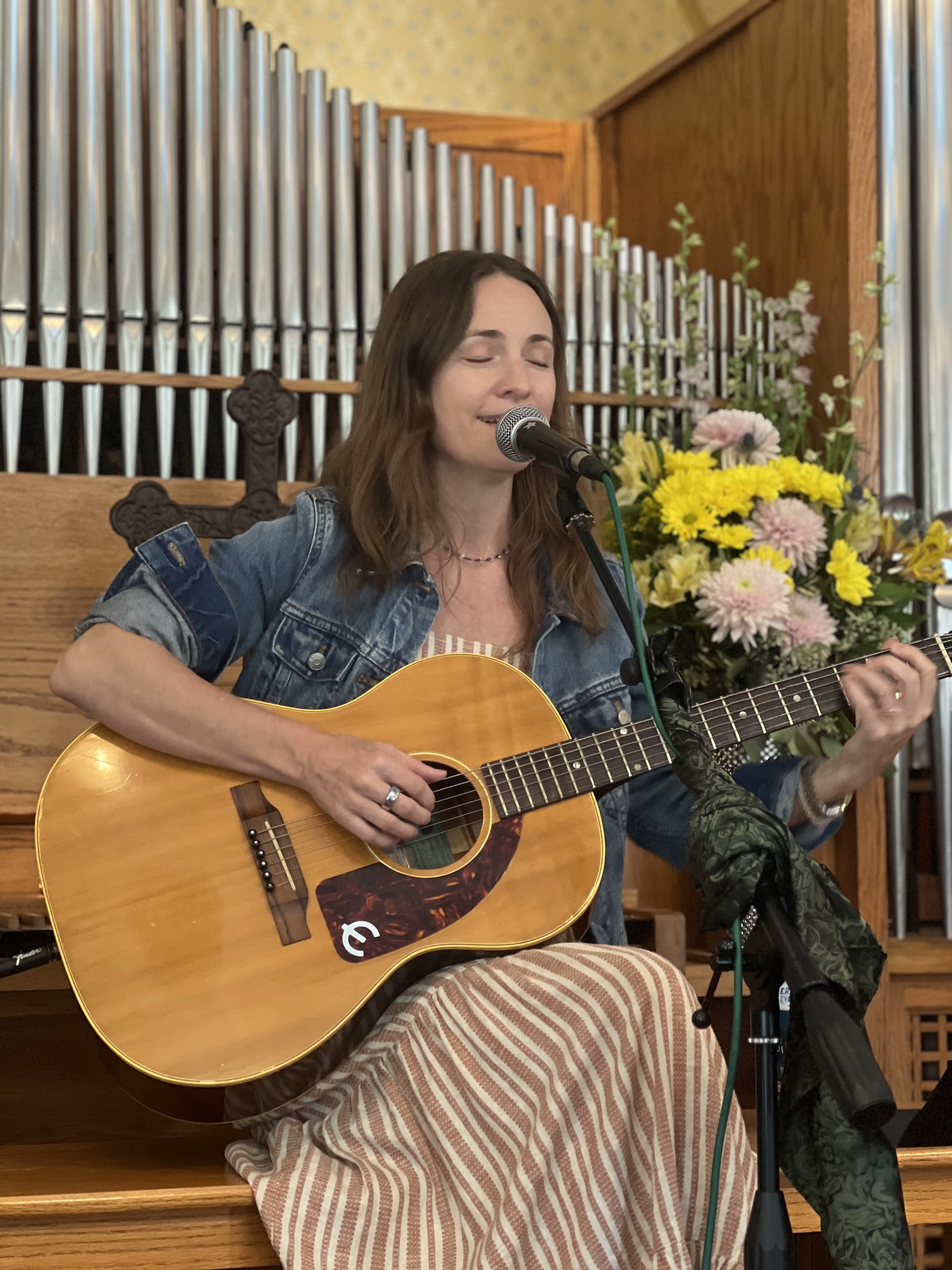 A woman with brown hair and a denim jacket playing an acoustic guitar and singing into a microphone, sitting in front of a floral arrangement and church organ pipes in a cozy indoor setting.