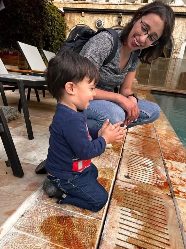A young boy kneeling on the floor, clapping and smiling at a metal grill on the ground, while a woman, possibly his mother, kneels nearby, laughing and watching him.