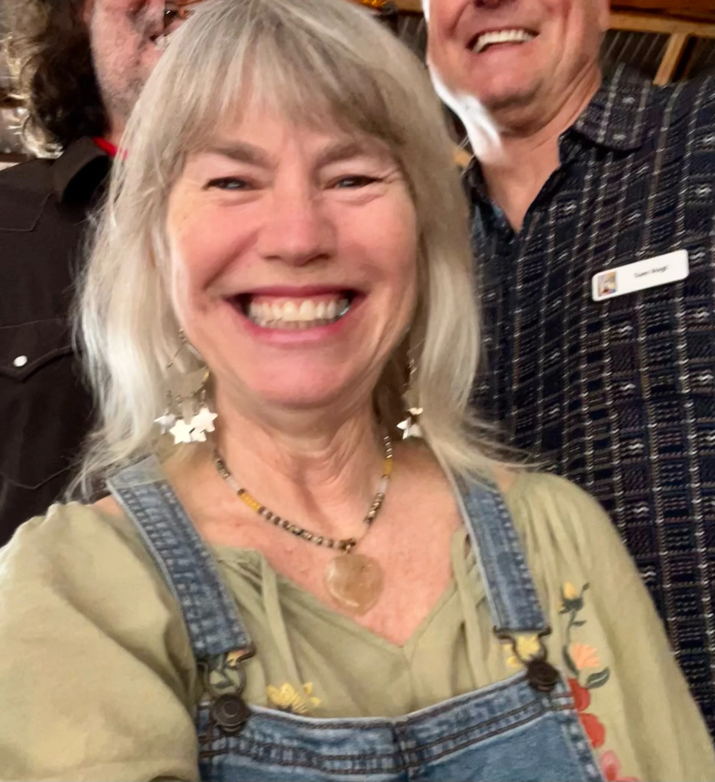 Close-up of smiling woman with blonde hair, wearing a green top, star-shaped earrings, necklace, and overalls, standing next to a man with a big smile, denim shirt, and a name tag.