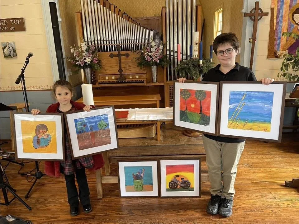A boy and a girl holding framed artwork inside a church, with a wooden piano, flowers, a cross, and religious paintings in the background.