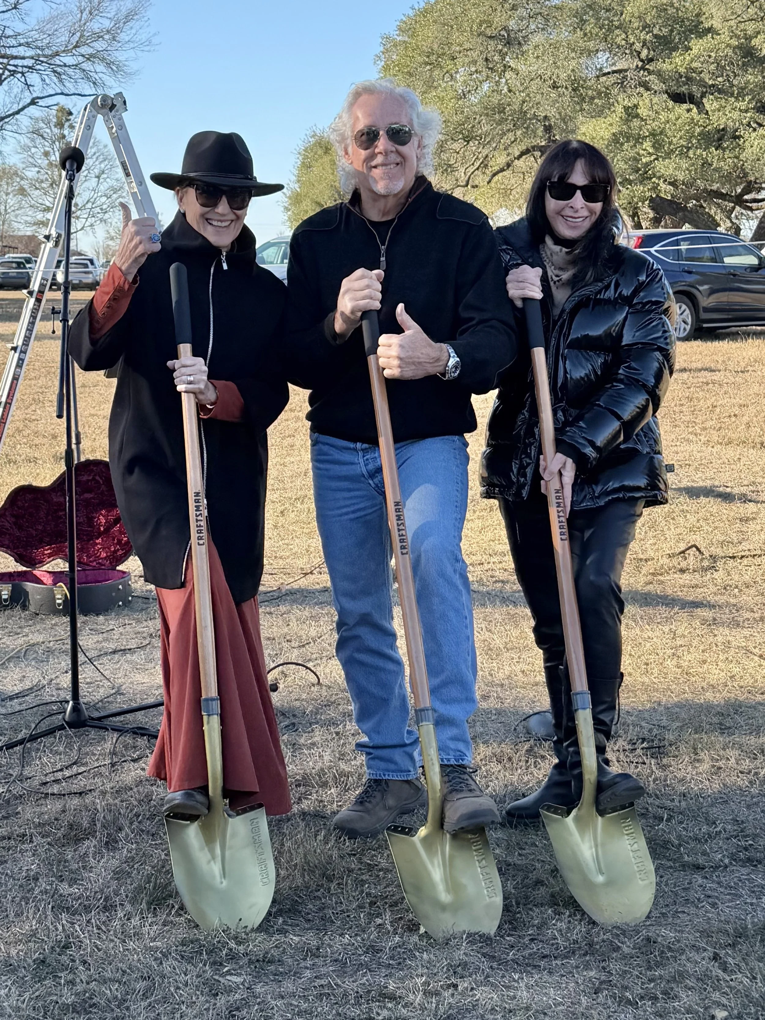 Three smiling adults wearing sunglasses and holding shovels outdoors on a grassy area with trees and parked cars in the background.