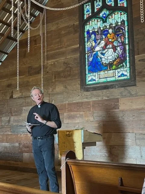 A man dressed as a priest standing inside a church next to a stained glass window, holding a book, and gesturing with his hand. The church has wooden walls, a pulpit, and hanging chains from the ceiling.