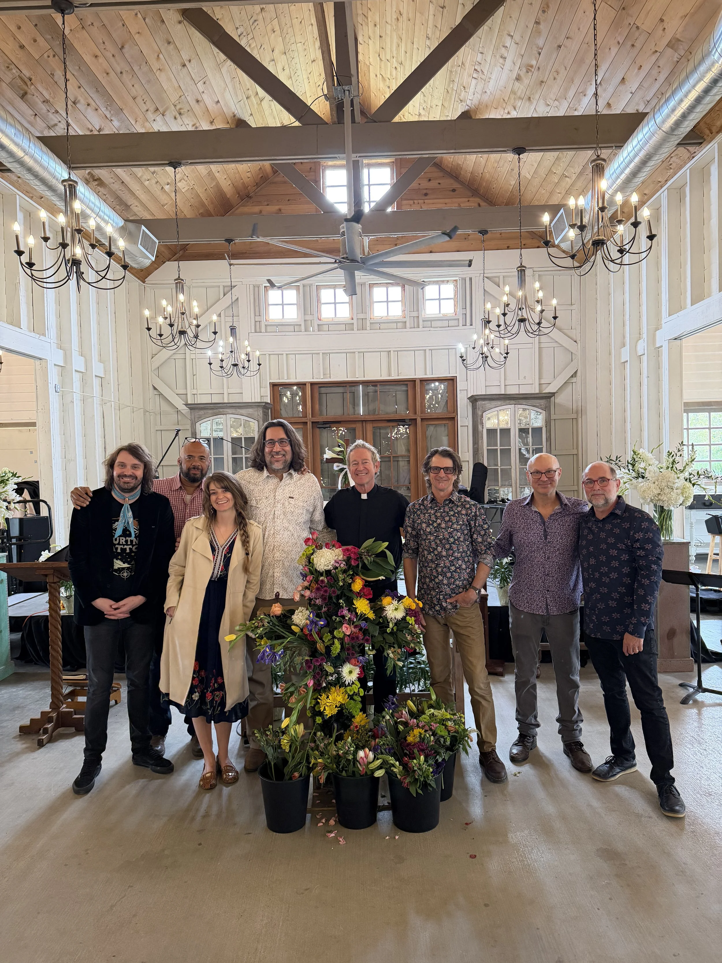 Group of nine people standing behind a table of colorful flowers in a rustic interior with wood-paneled walls, large windows, chandeliers, and exposed ductwork.