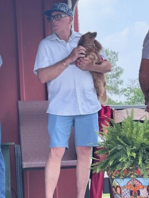 an older woman wearing glasses, a patterned cap, a white short-sleeve shirt, and blue shorts holding a small dog on a porch