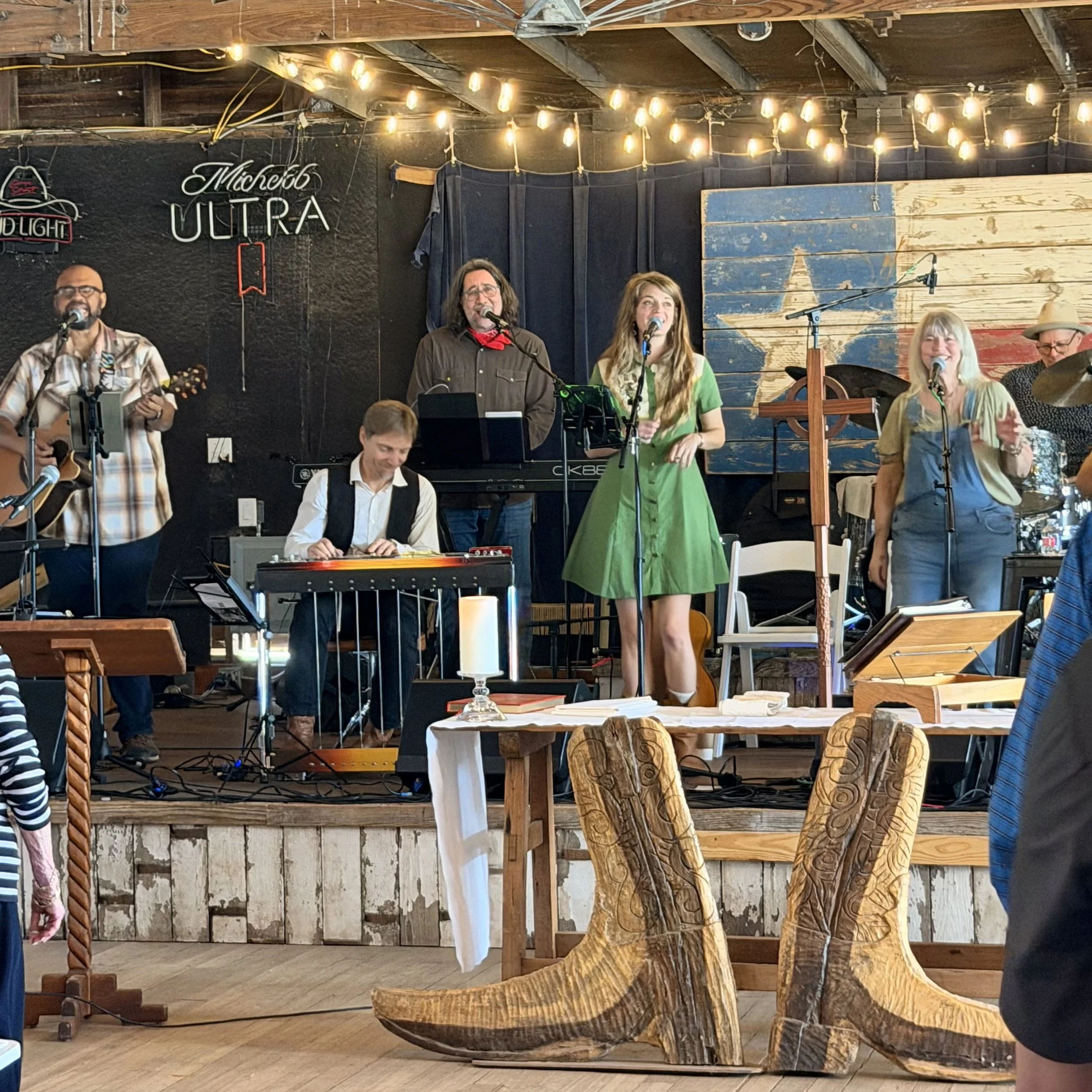 A group of musicians performing on a wooden stage with string lights overhead. There are five people visible, including a man playing guitar, a woman singing, and others playing instruments. The backdrop features a large American flag painted on wood