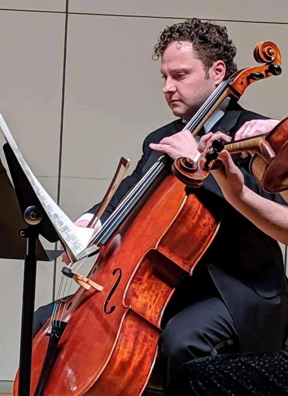 A man with curly hair playing a cello during a performance, with sheet music on a stand nearby.